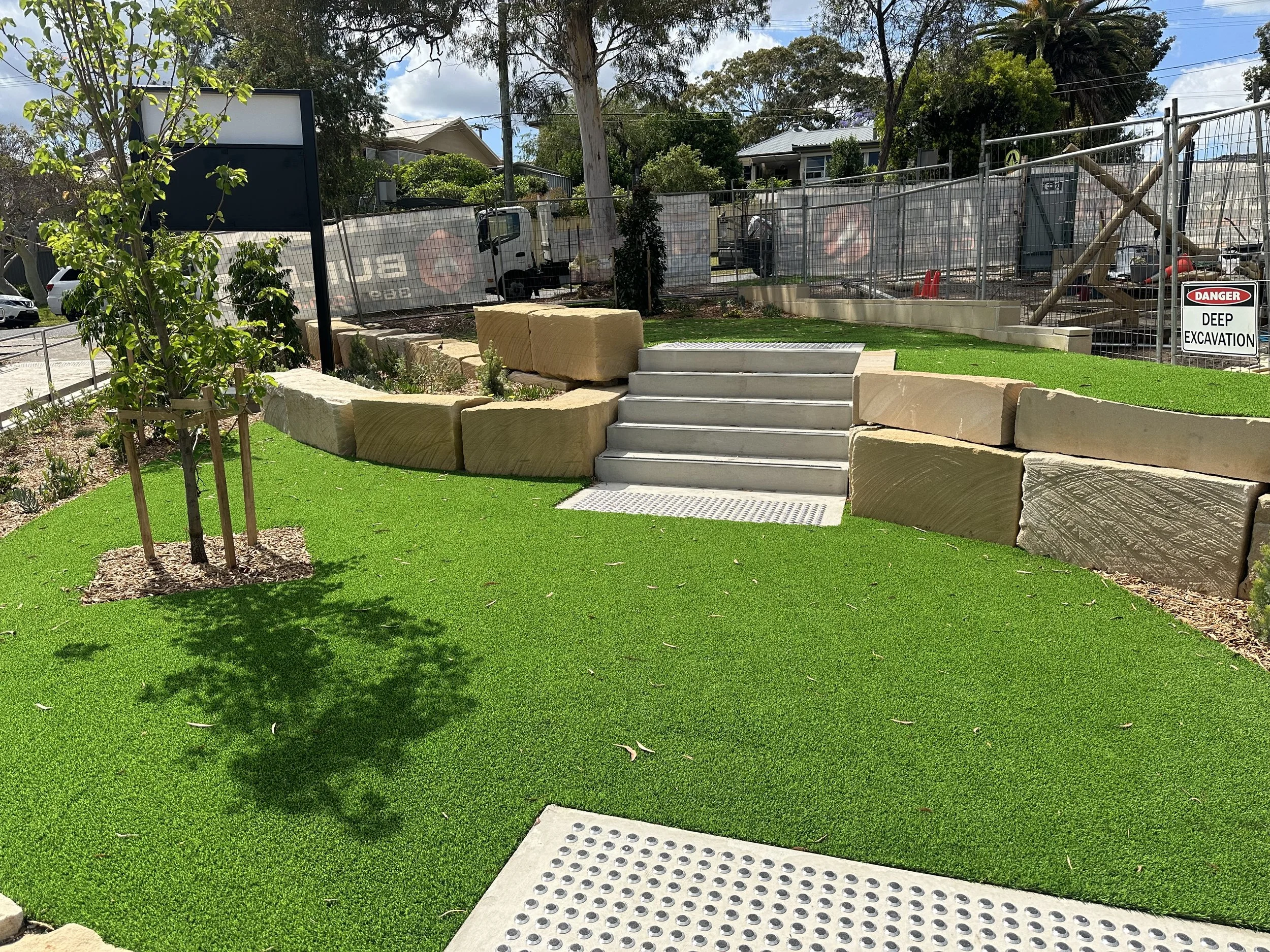 A landscaped area with artificial grass, small trees, stone border. Services by Greenscape Turfing Sydney