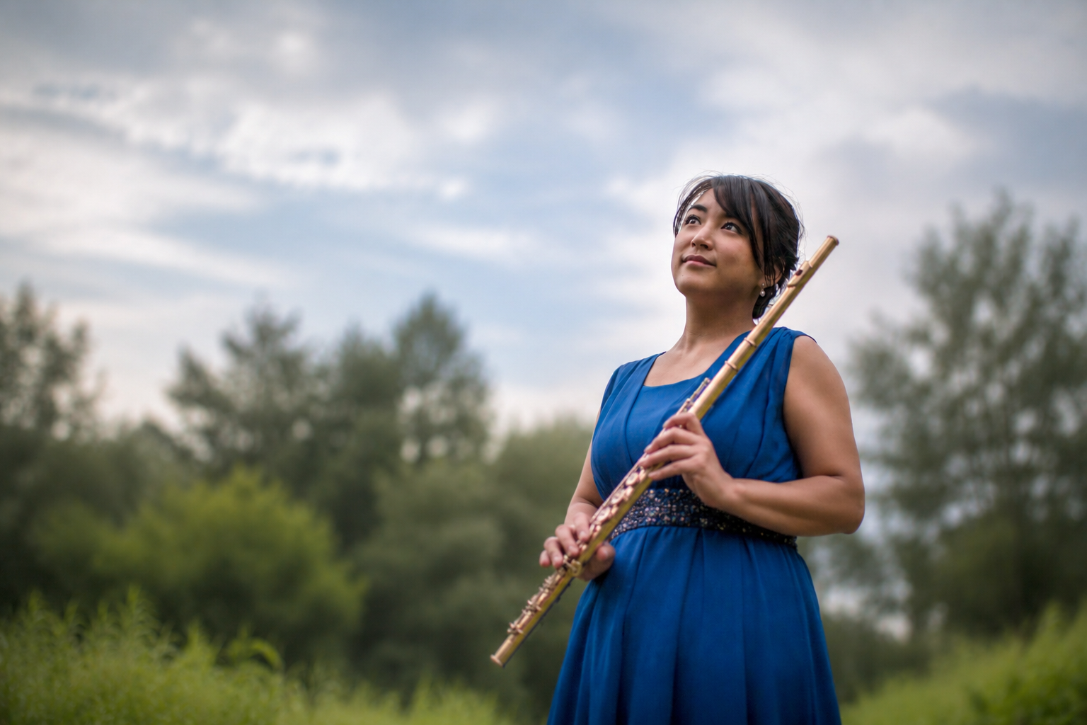 A woman in a blue dress holding a flute outdoors, looking up with a thoughtful expression, with trees and a cloudy sky in the background.