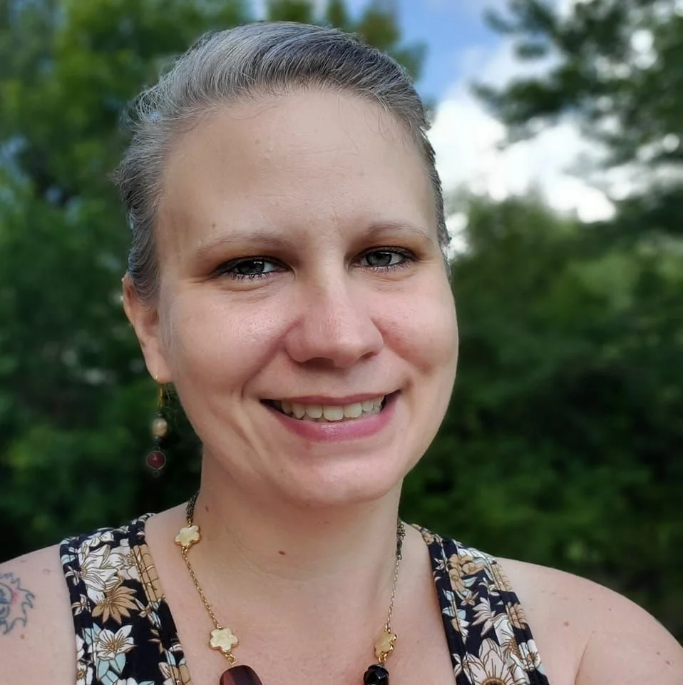 Une femme souriante avec des cheveux gris attachés, portant des bijoux en or et un haut à motifs floraux, en extérieur avec des arbres en arrière-plan.