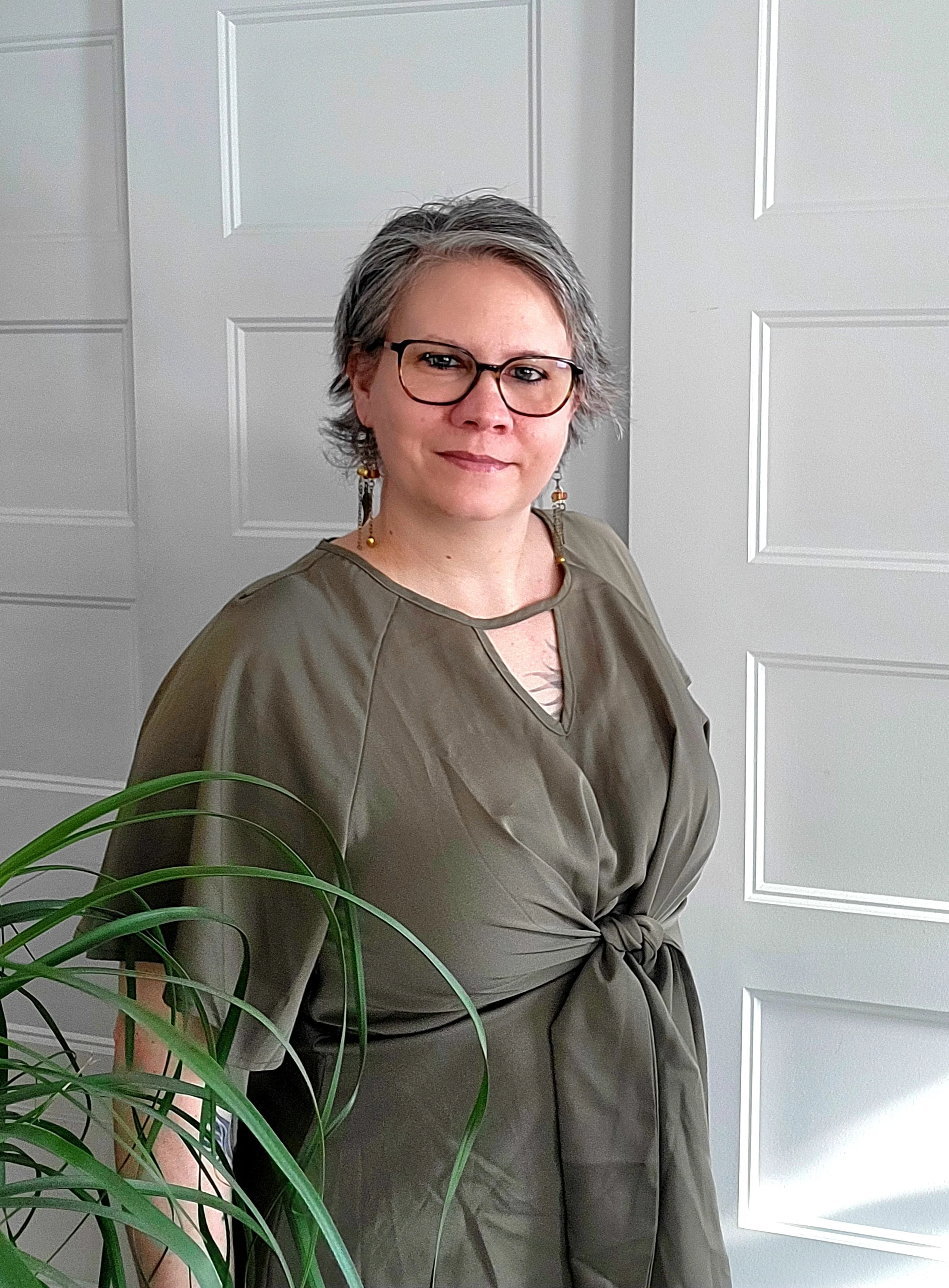Une femme avec des cheveux courts, porte des lunettes et des boucles d'oreilles, vêtue d'une chemise grise, se tenant devant une porte blanche. Un plant d'intérieur est partiellement visible en premier plan.