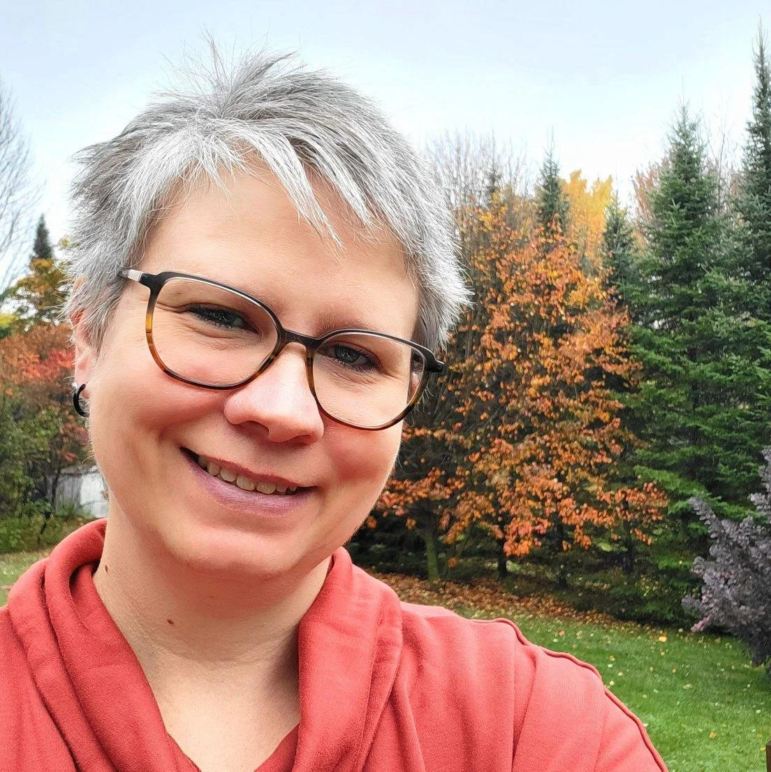 Une femme souriante portant des lunettes, prenant un selfie dans une jardin avec des arbres aux feuilles colorées d'automne.