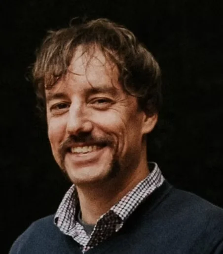Matt, with light brown curly hair and facial hair, wearing a dark blazer and a checkered shirt, standing against a dark background.