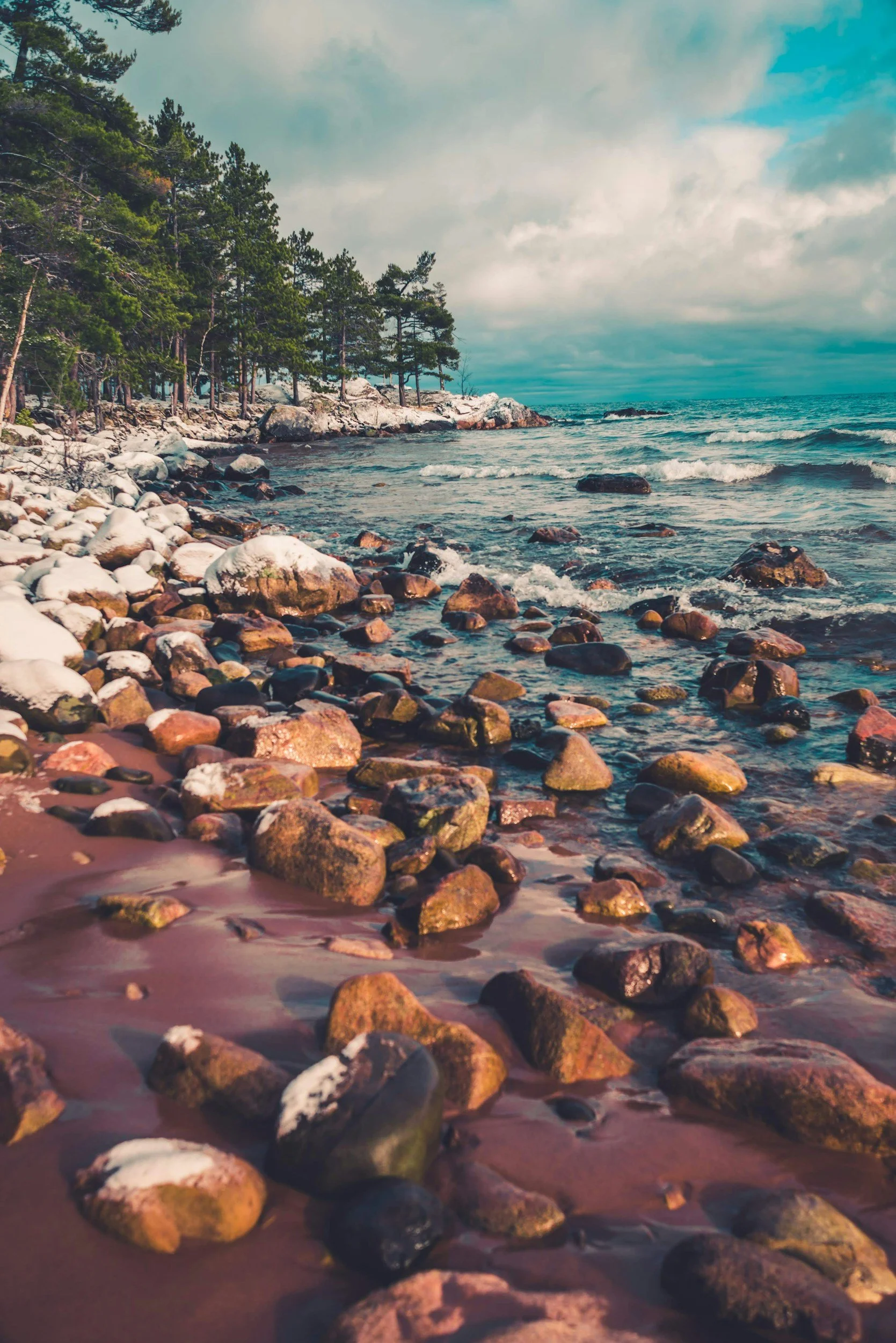 Rocks along a shoreline with trees on the left and water with small waves on the right, under a cloudy sky.