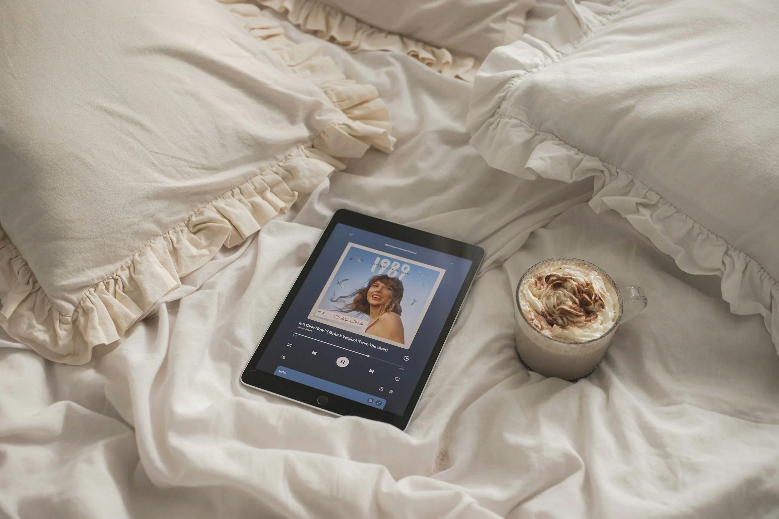 A bed with cream-colored ruffled bedding, a tablet displaying a music playlist, and a glass of iced coffee topped with whipped cream and chocolate drizzle.
