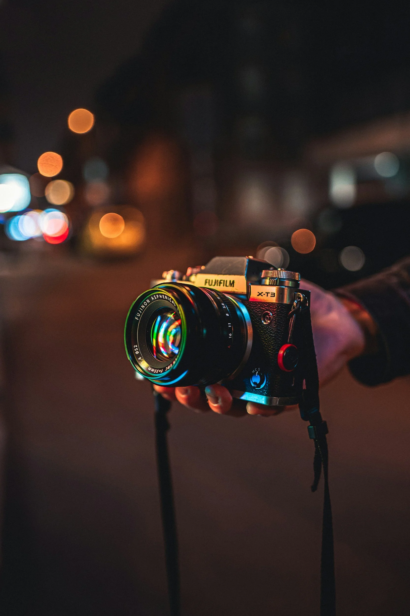 A hand holds a vintage camera with a reflective lens, capturing city lights bokeh in the background. The scene conveys a nostalgic, artistic mood.