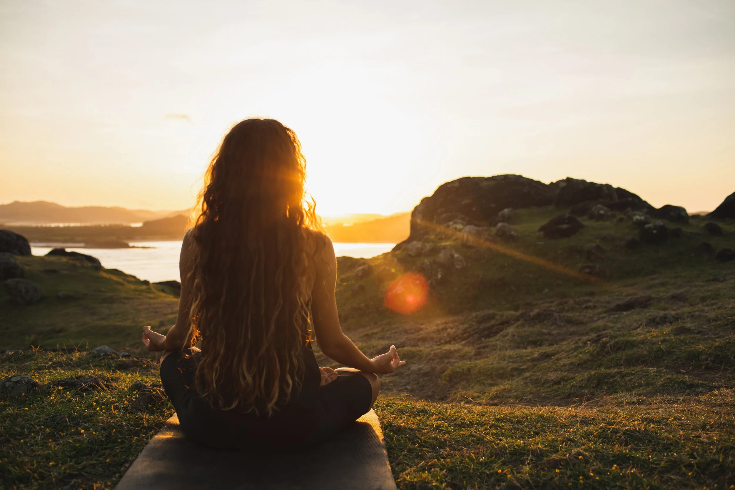 A person with long hair sits in a meditative pose on a grassy hill at sunset, facing the sun. The scene is peaceful and serene, with soft golden light.