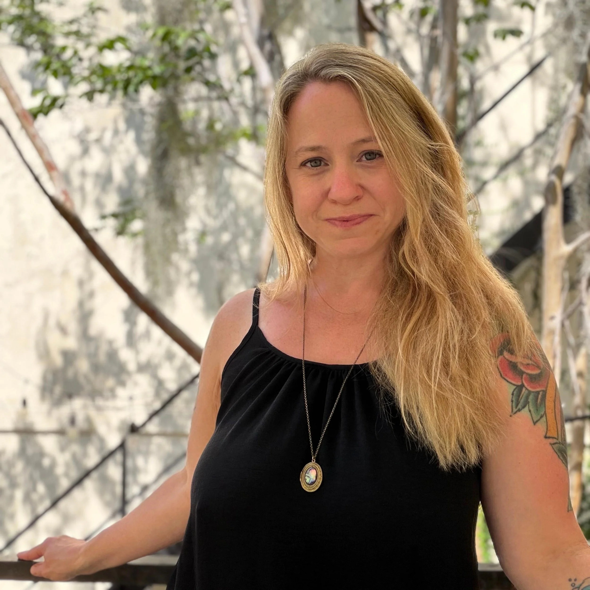 Portrait of Sasha Trickett with long blonde hair, wearing a black tank top and a pendant necklace, standing outdoors in natural light.
