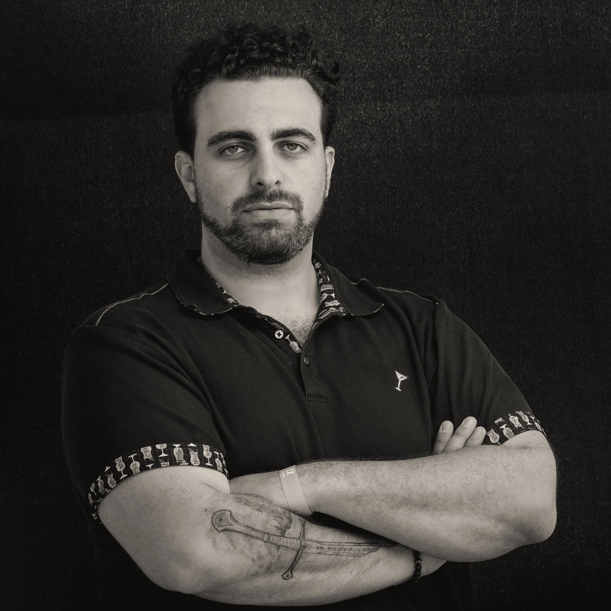 Black and white photo of a young man with styled hair, beard, and tattoos on his arm, wearing a black polo shirt with patterned cuffs, standing in front of a textured surface.