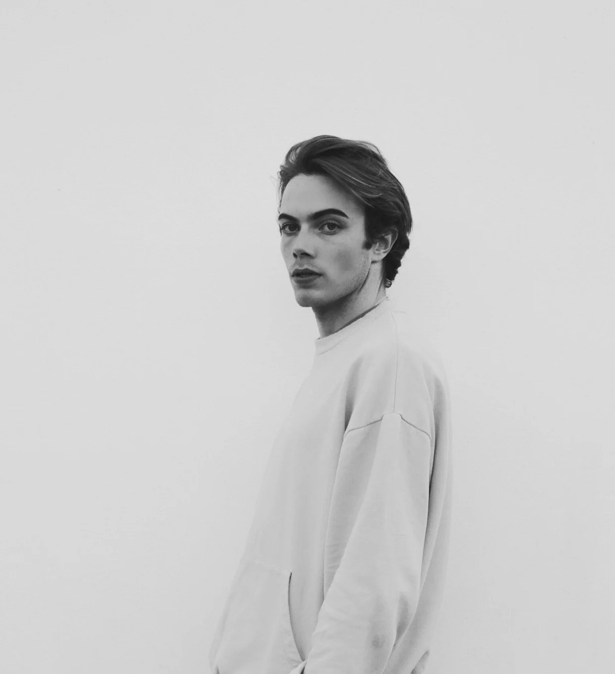 Black and white photo of a young man with dark hair and defined eyebrows, wearing a light-colored sweatshirt, standing against a plain white wall, looking at the camera with a serious expression.