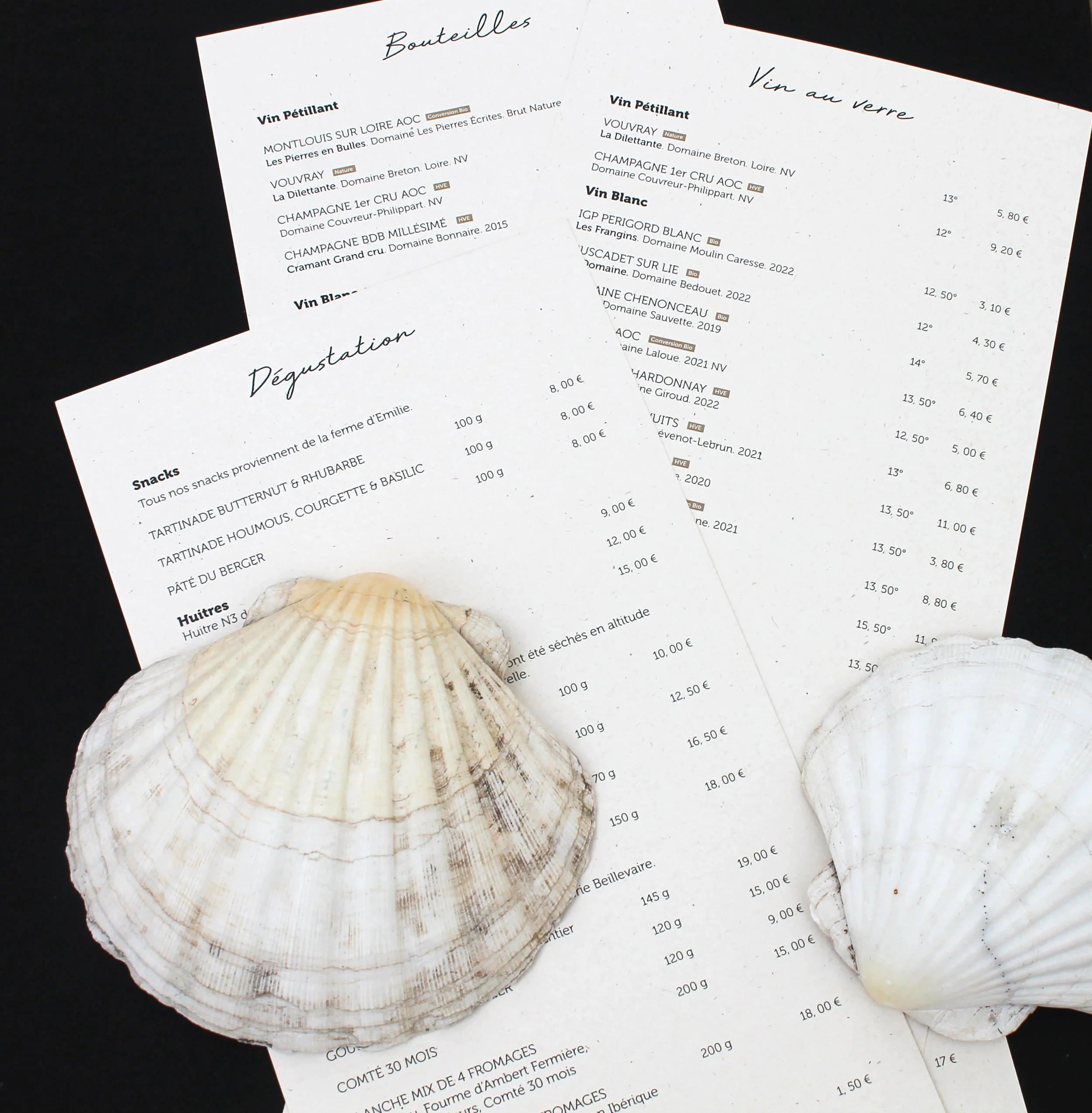Deux menus de restaurant ou de vin affichés d'une main, avec coquilles Saint-Jacques en premier plan, cassées et ouvertes, devant un fond noir.
