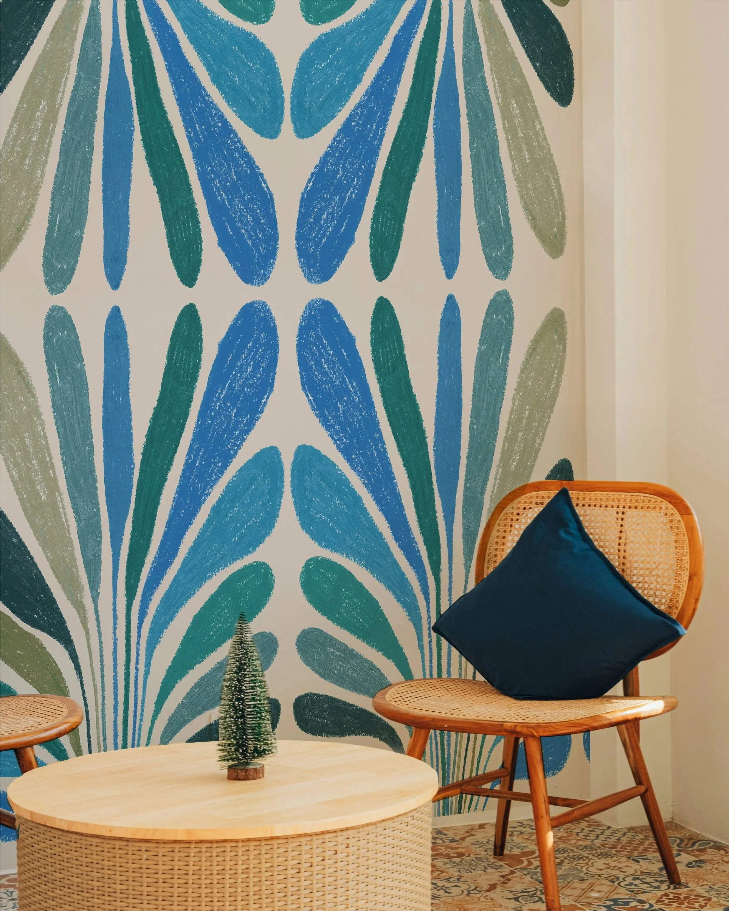 Un coin salon avec deux chaises en bois et un canapé avec un coussin bleu foncé, décoré d'une petite sapin en haut d'une table en bois et d'un mur avec un grand motif floral bleu et vert.
