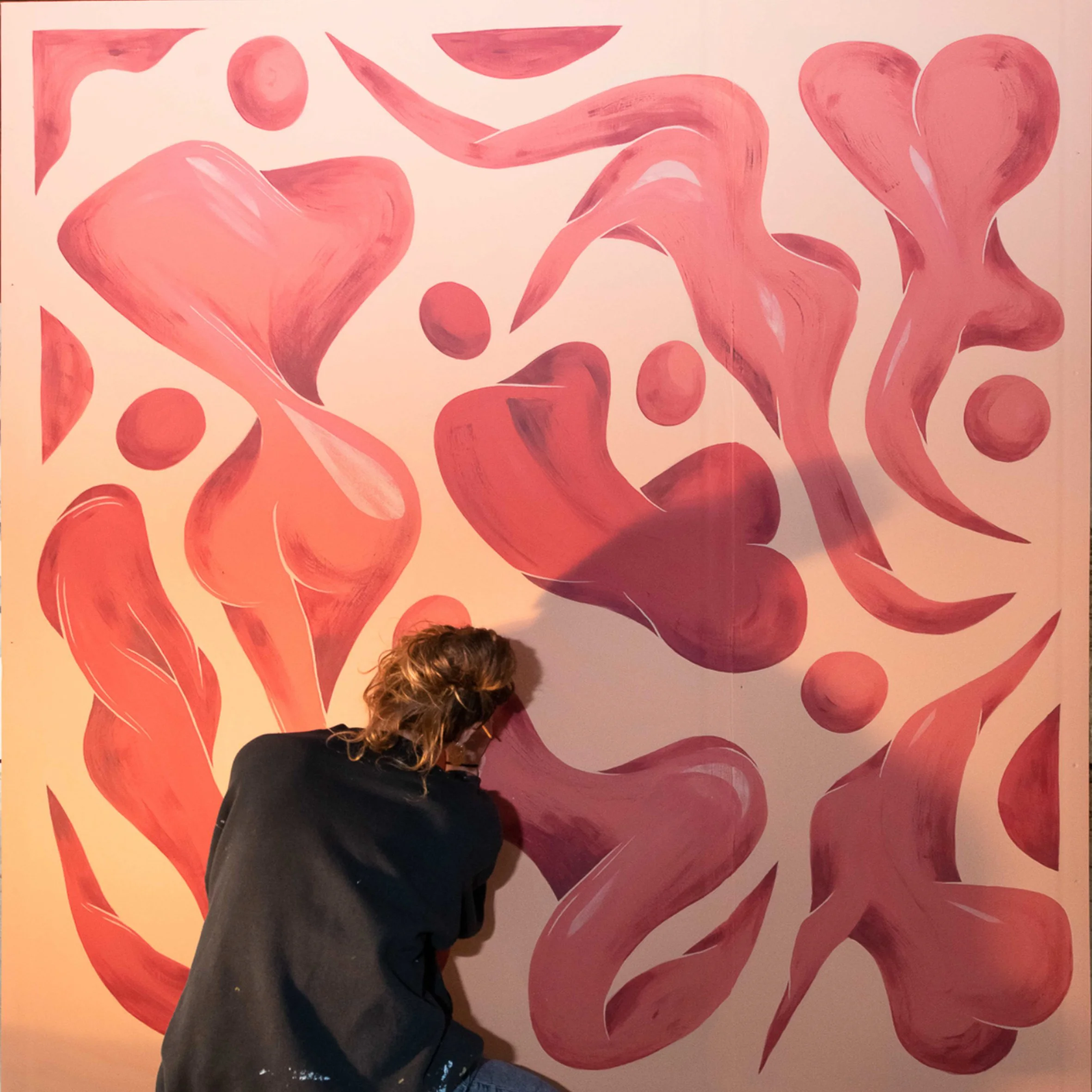 Une personne avec des cheveux bruns et vêtue d'une veste noire, observant un mur avec une peinture abstraite composée de formes rouges.