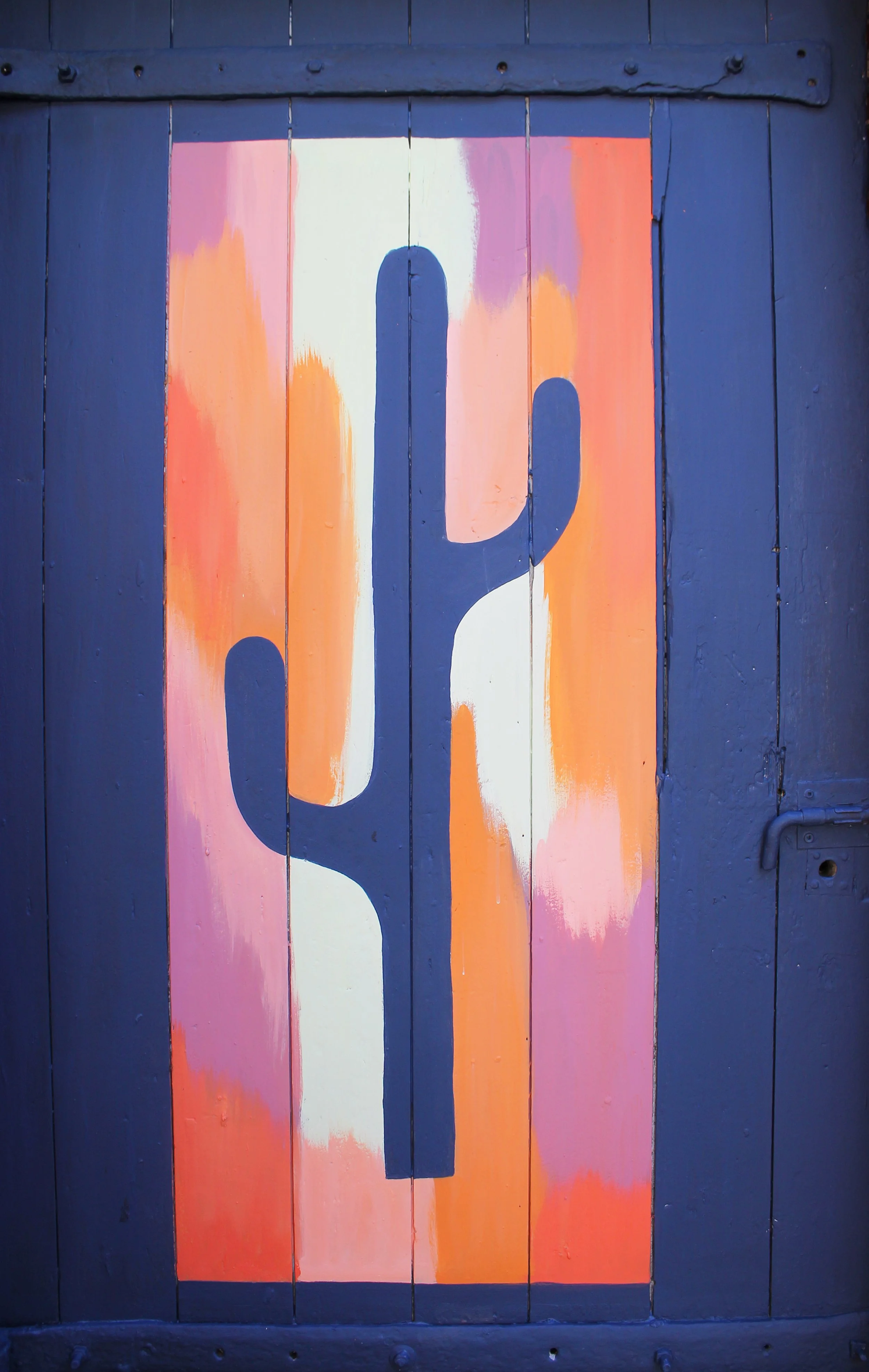 Une peinture abstraite représentant un cactus stylisé sur une porte en bois peinte de couleurs chaudes comme le rose, l'orange et le blanc, avec un fond bleu foncé.