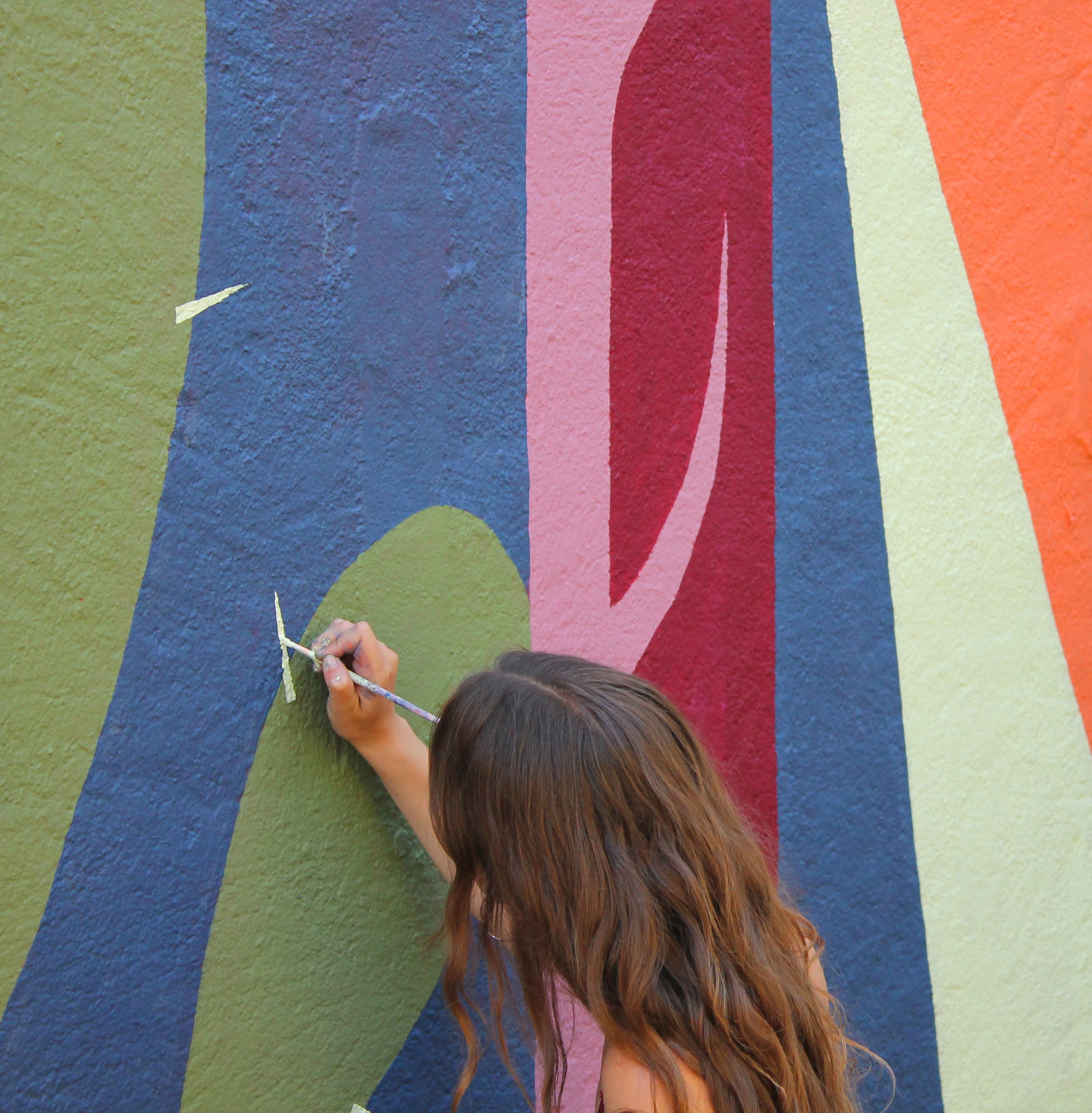 Une personne en train de peindre un mur en couleurs vives avec des formes abstraites. La personne a les cheveux bruns longs.