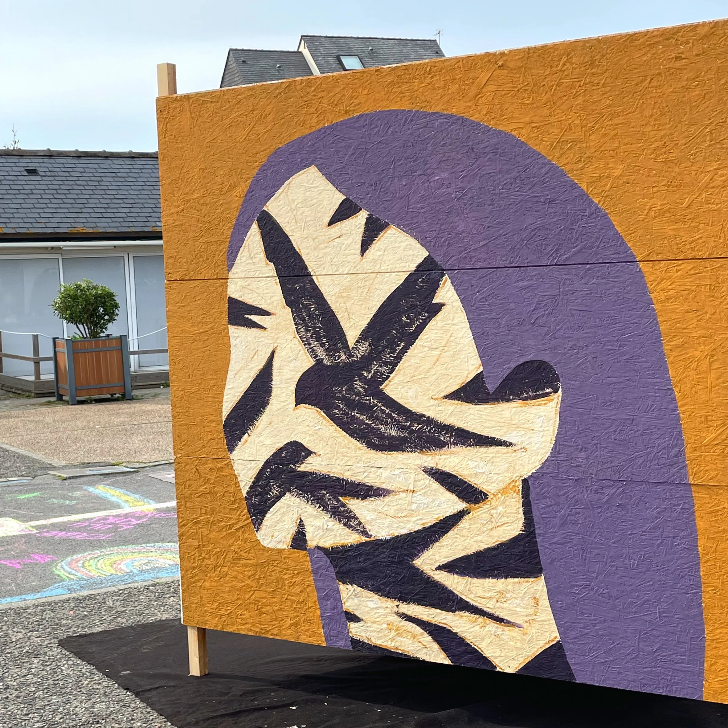 Une œuvre d'art murale colorée représentant un visage humain en style abstrait, avec des couleurs vives de violet, beige, noir et orange, installée dans une zone urbaine avec un bâtiment résidentiel en arrière-plan, sur un support en bois avec du gravier au sol.
