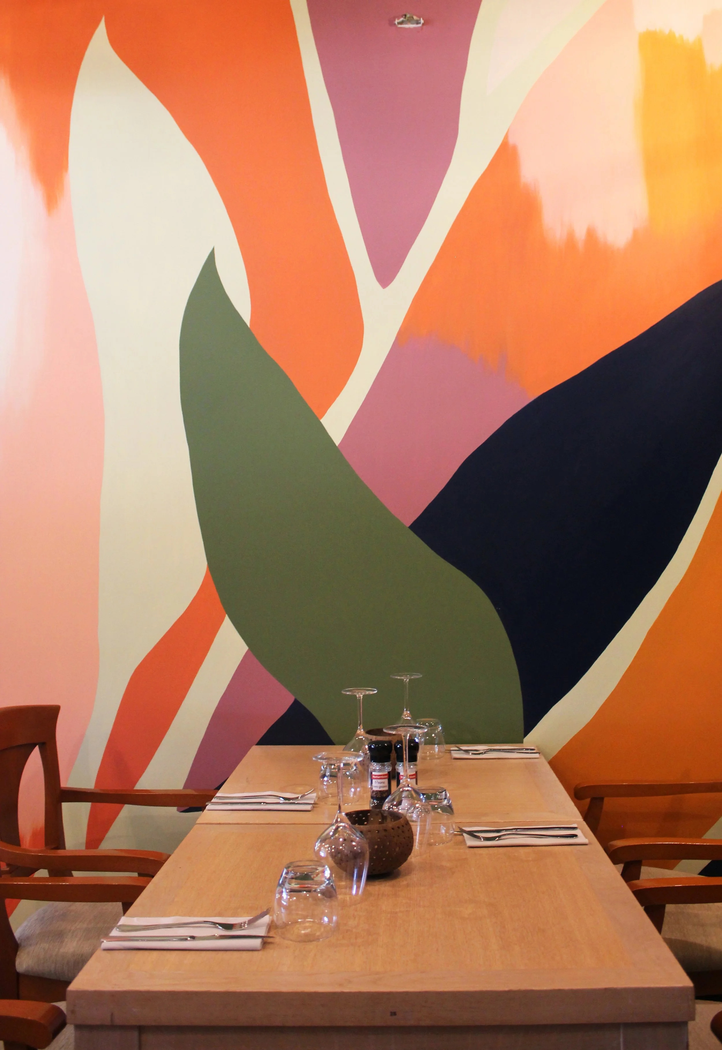 Salle à manger avec une grande table en bois, quatre verres à vin, couverts et assiettes, mur peint avec un motif abstrait aux couleurs orange, rose, vert et noir.