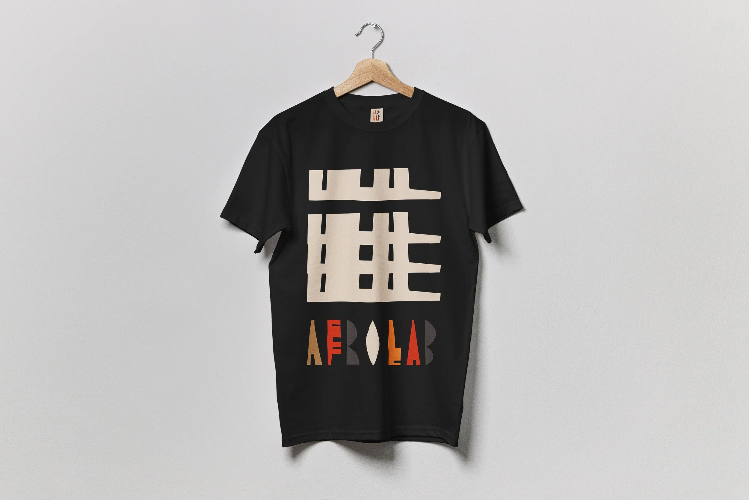 Un t-shirt noir avec un motif géométrique blanc et des lettres colorées rouges, orange, et beige, suspendu à un cintre en bois sur un mur blanc.