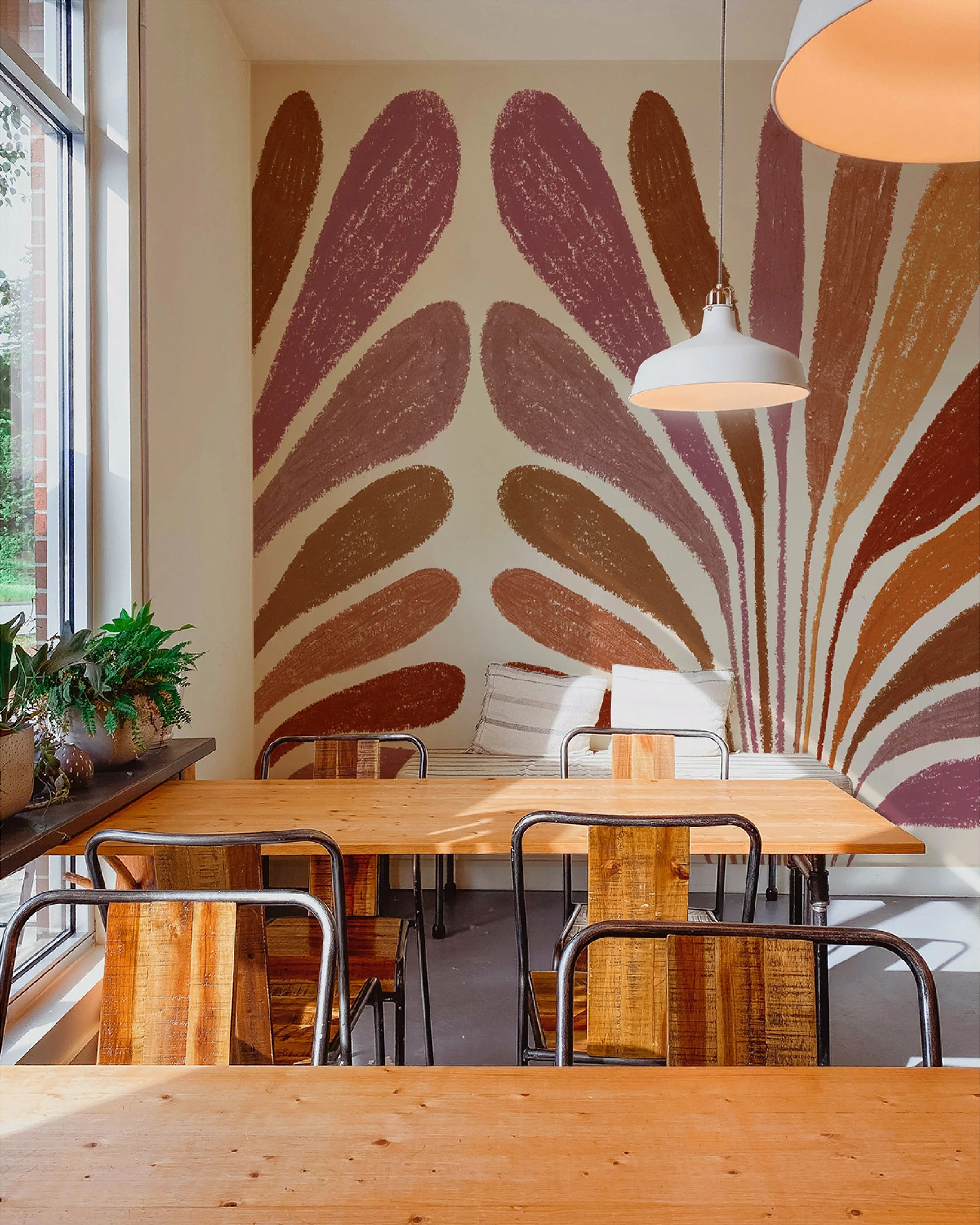 Une salle à manger avec une grande table en bois, des chaises en métal, un mur avec une fresque de motifs abstraits colorés, une fenêtre avec des plantes à gauche, et deux lampes suspendues à plafond. Il y a un coussin blanc avec des rayures sur un canapé ou une chaise en arrière-plan.