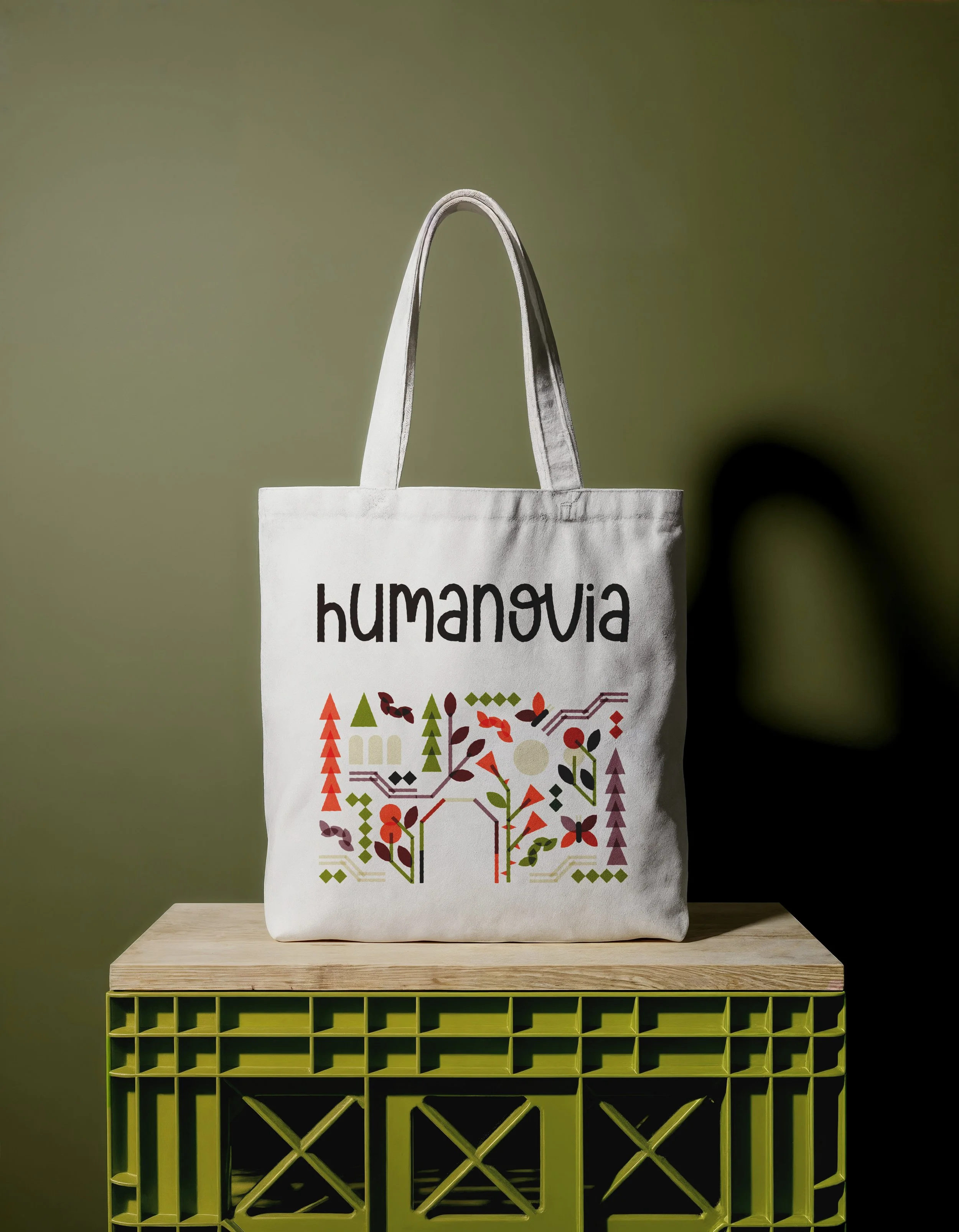 Sac en toile blanc avec le texte 'humaniovia' écrit en noir, posé sur une étagère en bois, avec une grille verte en dessous et un fond vert flou.
