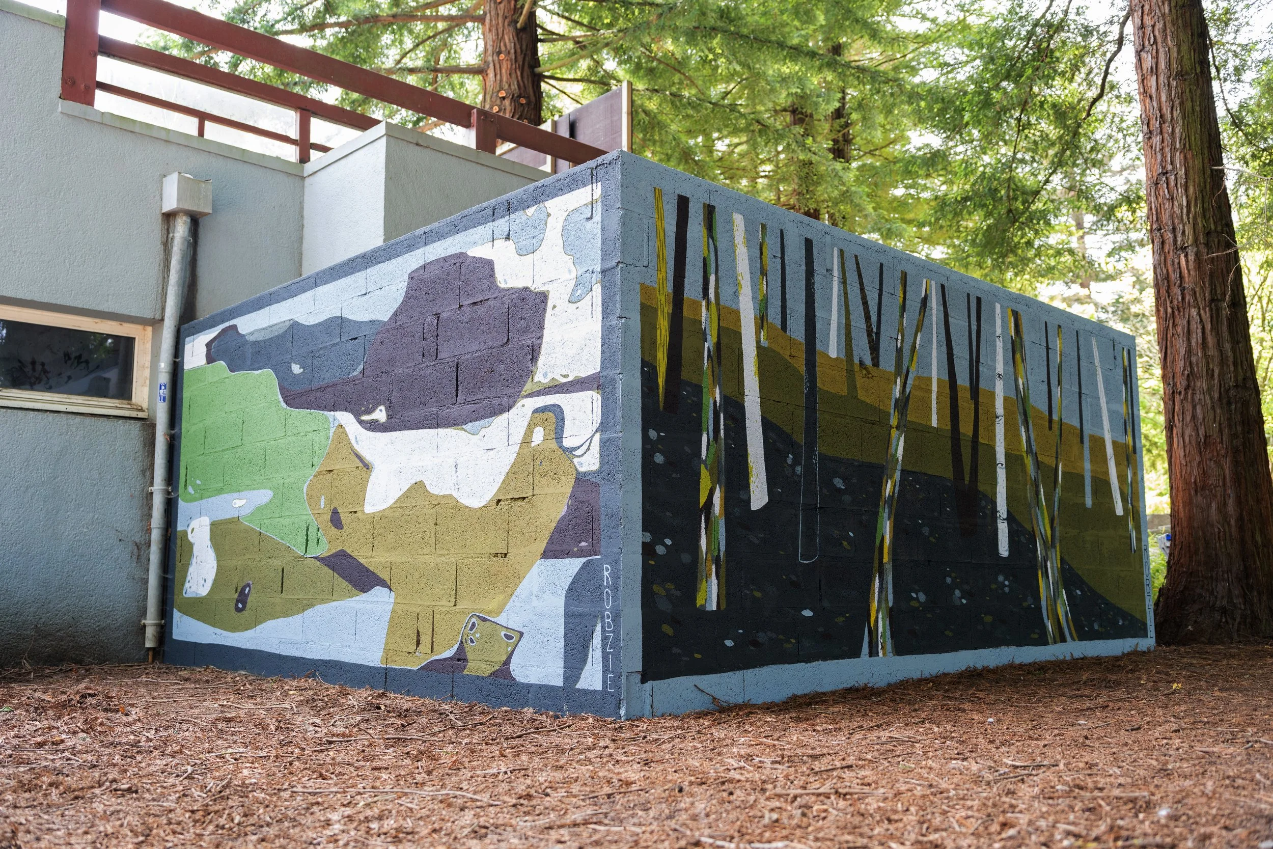 Murale en plein air représentant un paysage naturel avec des montagnes et des arbres, située à l'angle d'un bâtiment en béton peint, entourée d'arbres dans une forêt.