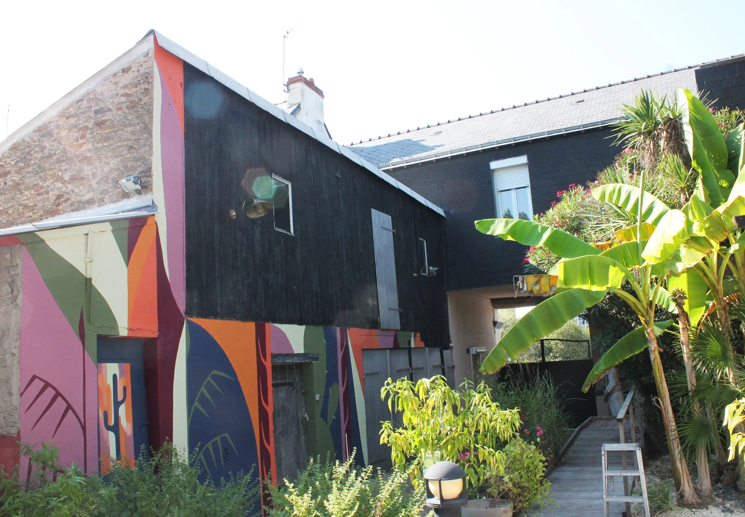 Facade d'une maison avec un mur coloré de motifs abstraits, entourée de végétation verte et d'un chemin en bois.