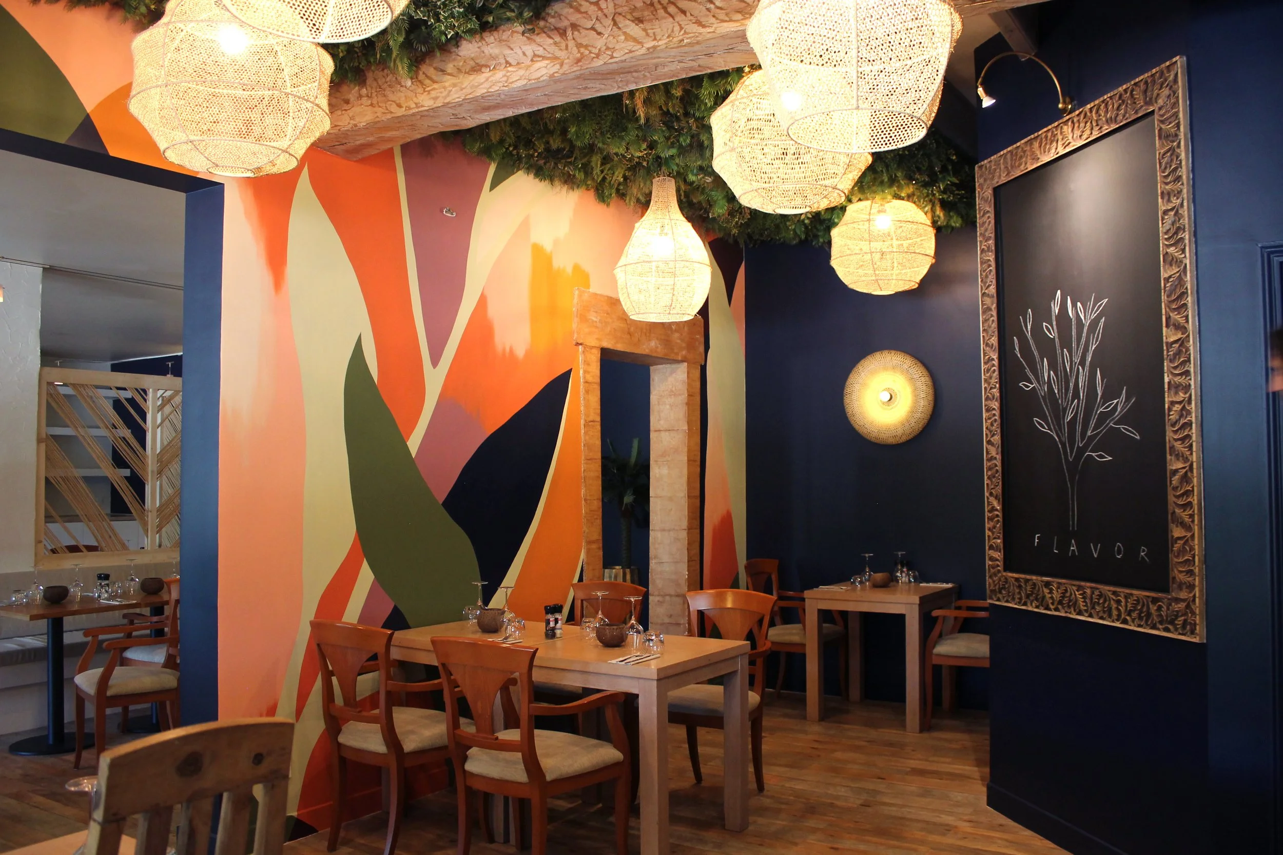 Intérieur d'un restaurant avec des tables en bois, des chaises, des murs colorés avec un mur peint multicolore, un grand miroir encadré en bois et des lampes en osier suspendues. Décorations vertes au plafond.