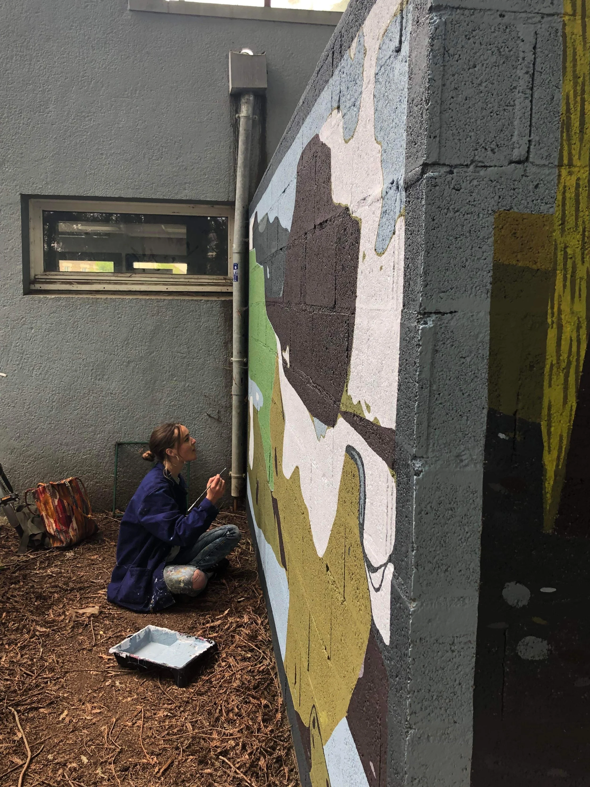 Une jeune femme en vêtement bleu peint une fresque murale en extérieur, au sol des brosses et une boîte de peinture, un bâtiment en arrière-plan, le mur est gris