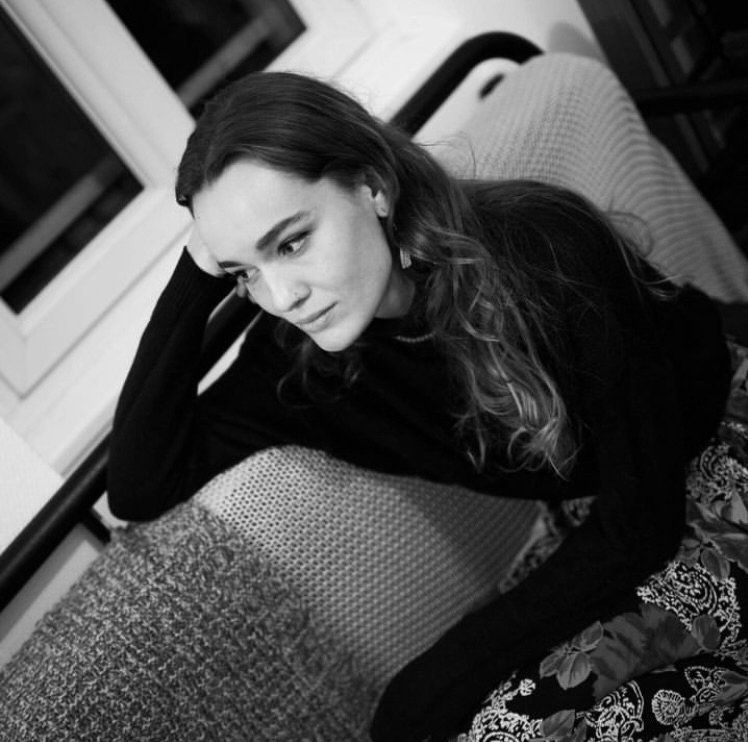 Une femme assise sur un canapé, en train de regarder pensivement vers la gauche, avec des cheveux longs et ondulés, portant un pull noir.