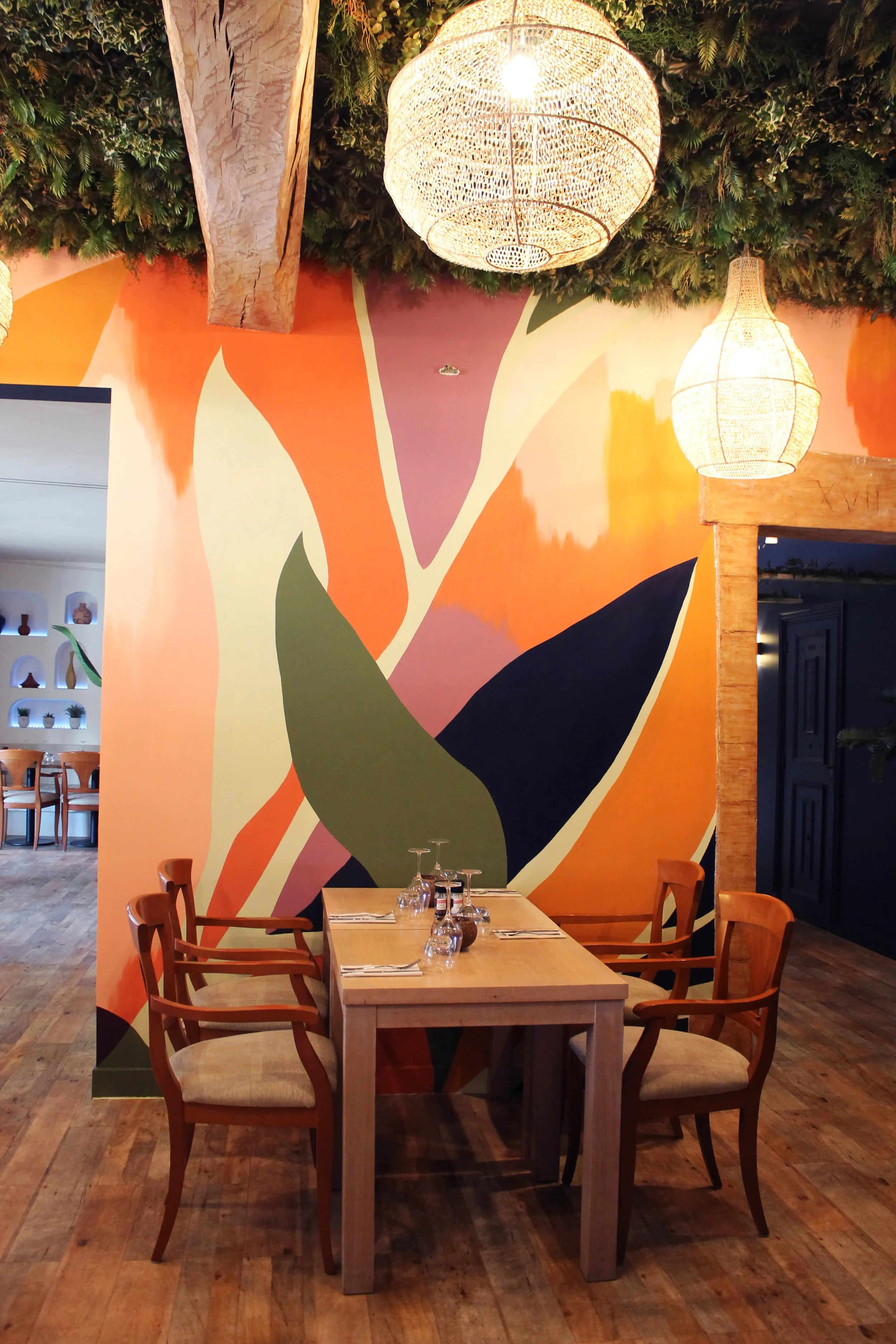 Intérieur d'un restaurant avec un mur peint aux couleurs vives, comprenant une grande fresque abstraite, des luminaires suspendus en forme de paniers en osier, des tables en bois avec des verres et des couverts, et un sol en bois. Il y a aussi des plantes vertes suspendues au plafond.
