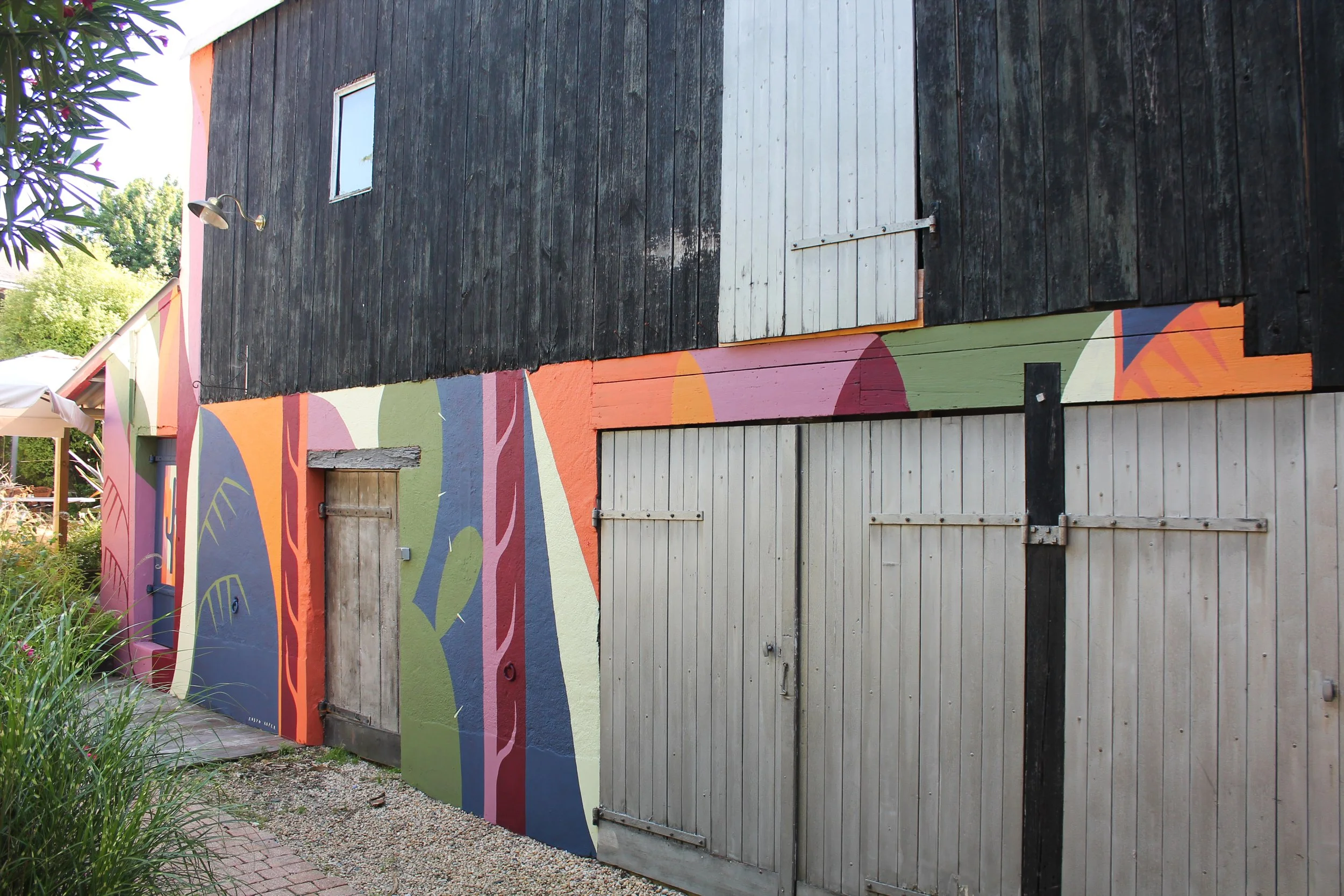 Façade d'un bâtiment avec une partie en bois peint en noir et une partie en bois peint en blanc, avec un mur orné d'une fresque colorée représentant des formes géométriques et des arbres stylisés.