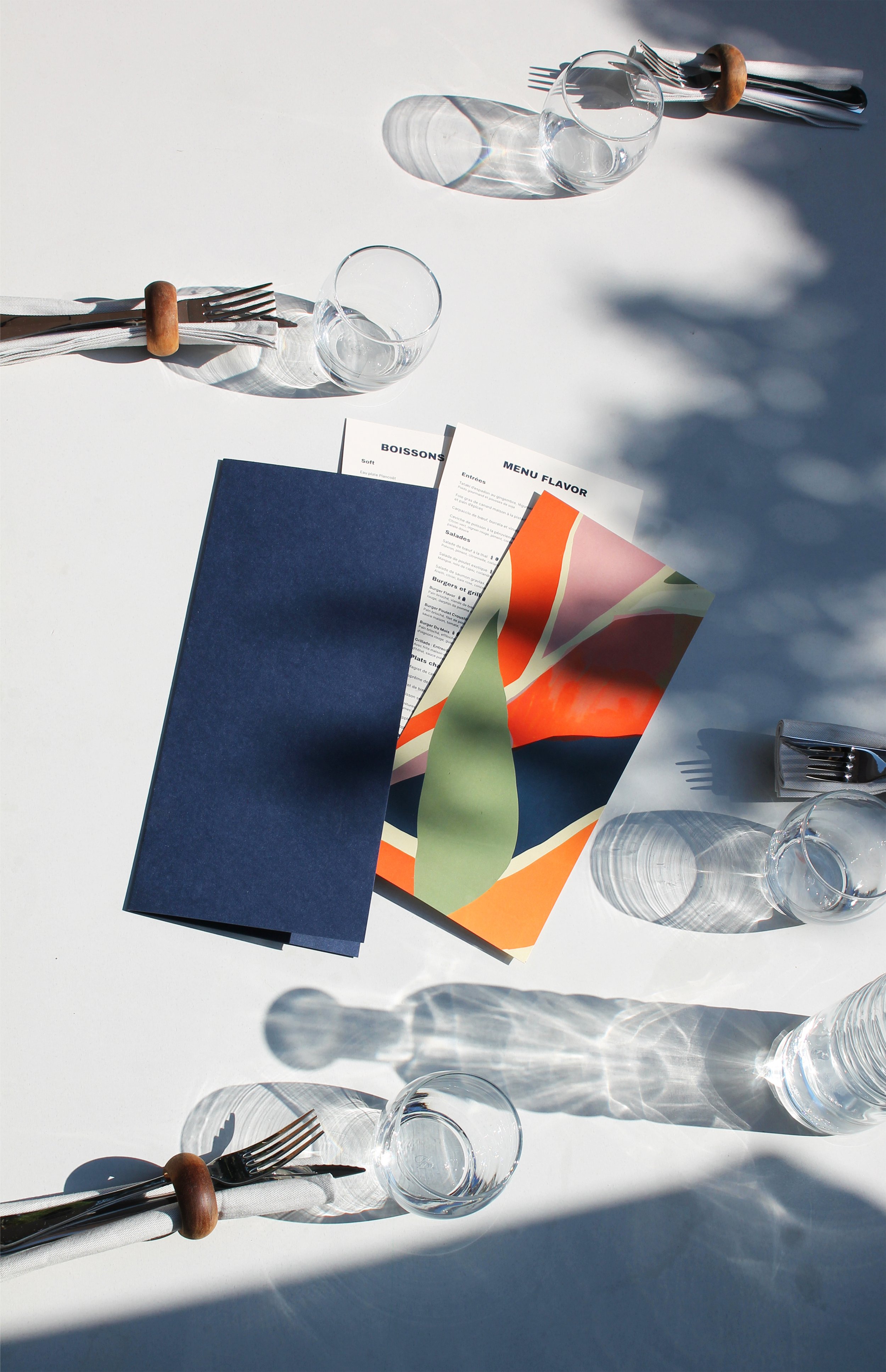 Une table dressée avec des verres, couverts, menu, et une nappe blanche sous une lumière naturelle créant des ombres.