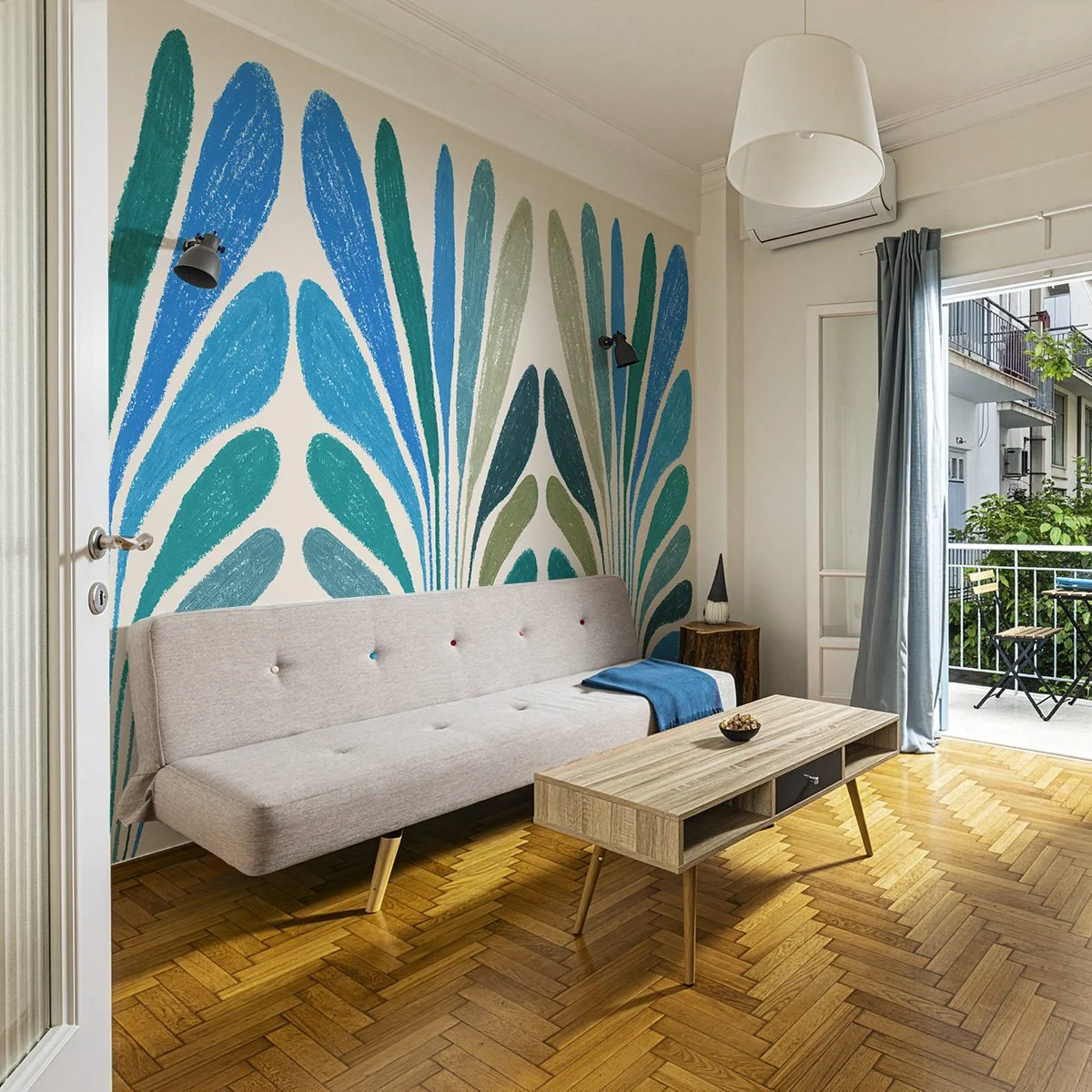 Salon avec un canapé beige, une table en bois, mur avec une fresque de feuilles bleues et vertes, une porte-fenêtre donnant sur un balcon avec des chaises, un lampadaire blanc, et un climatiseur mural.