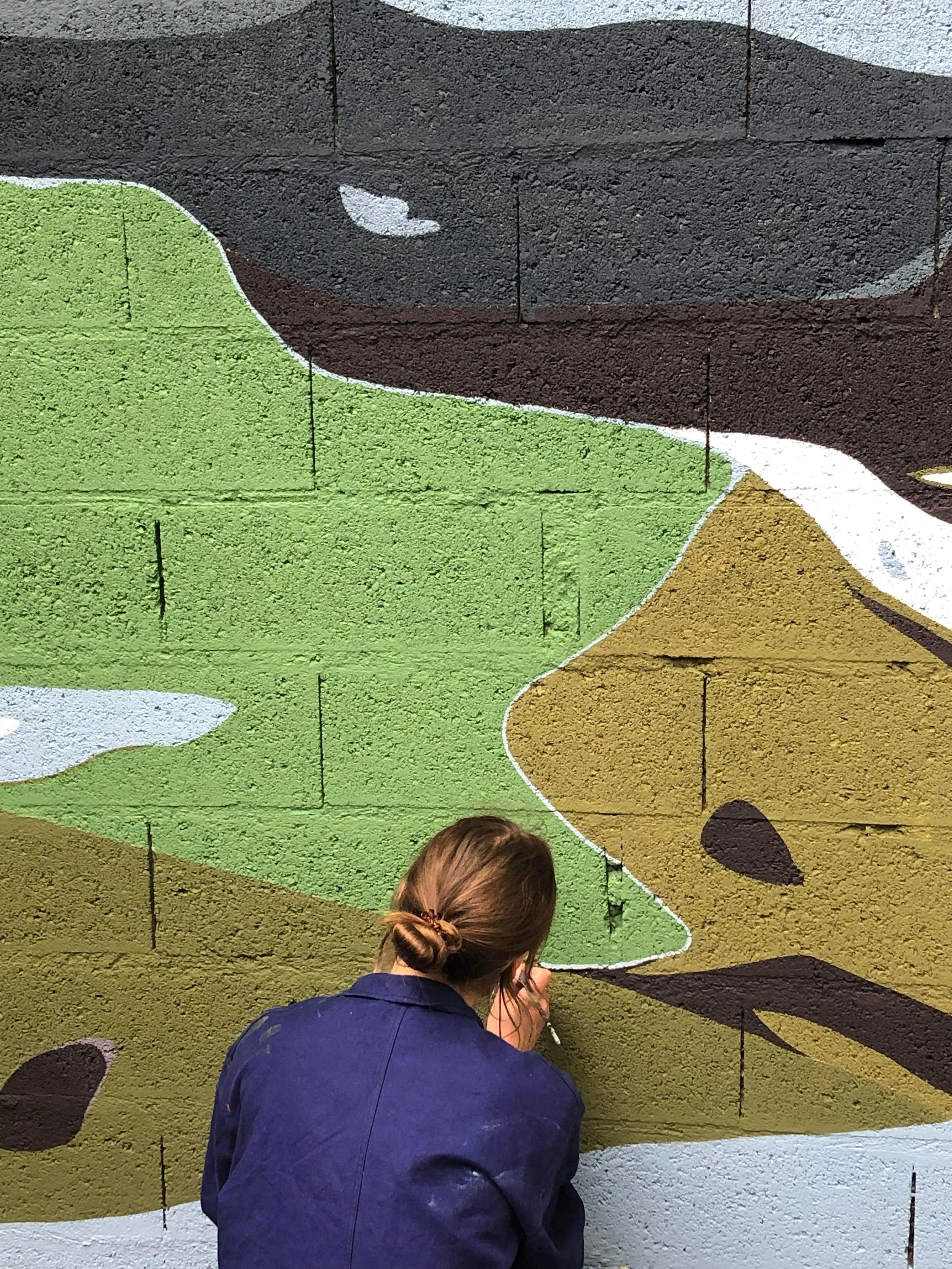 Une femme en train de peindre une fresque murale représentant des oiseaux colorés sur un mur en briques.