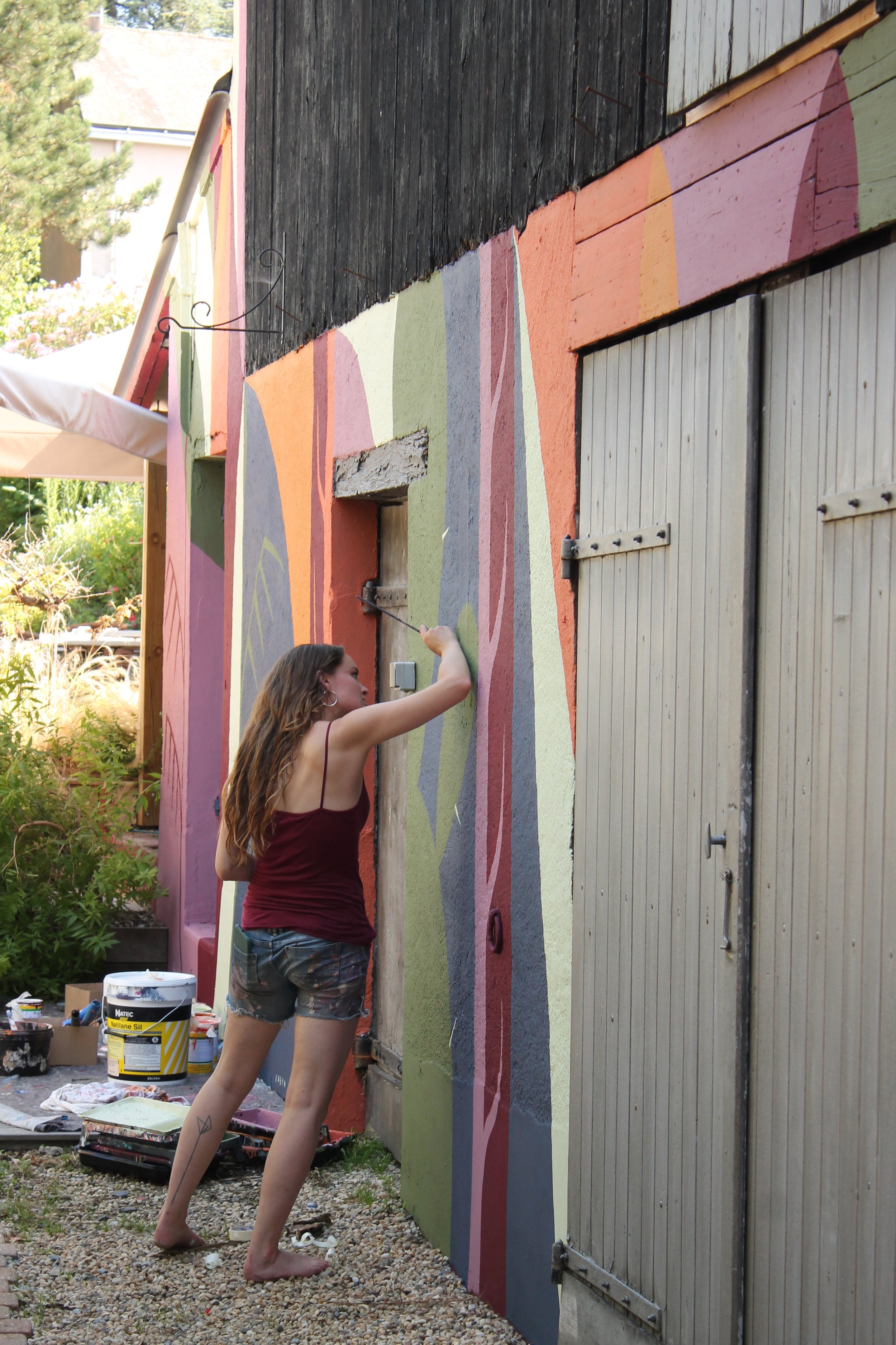 Une femme en jeans et haut sans manches peint un mur extérieur avec des couleurs pastel et des formes abstraites, à côté d'une porte en bois.