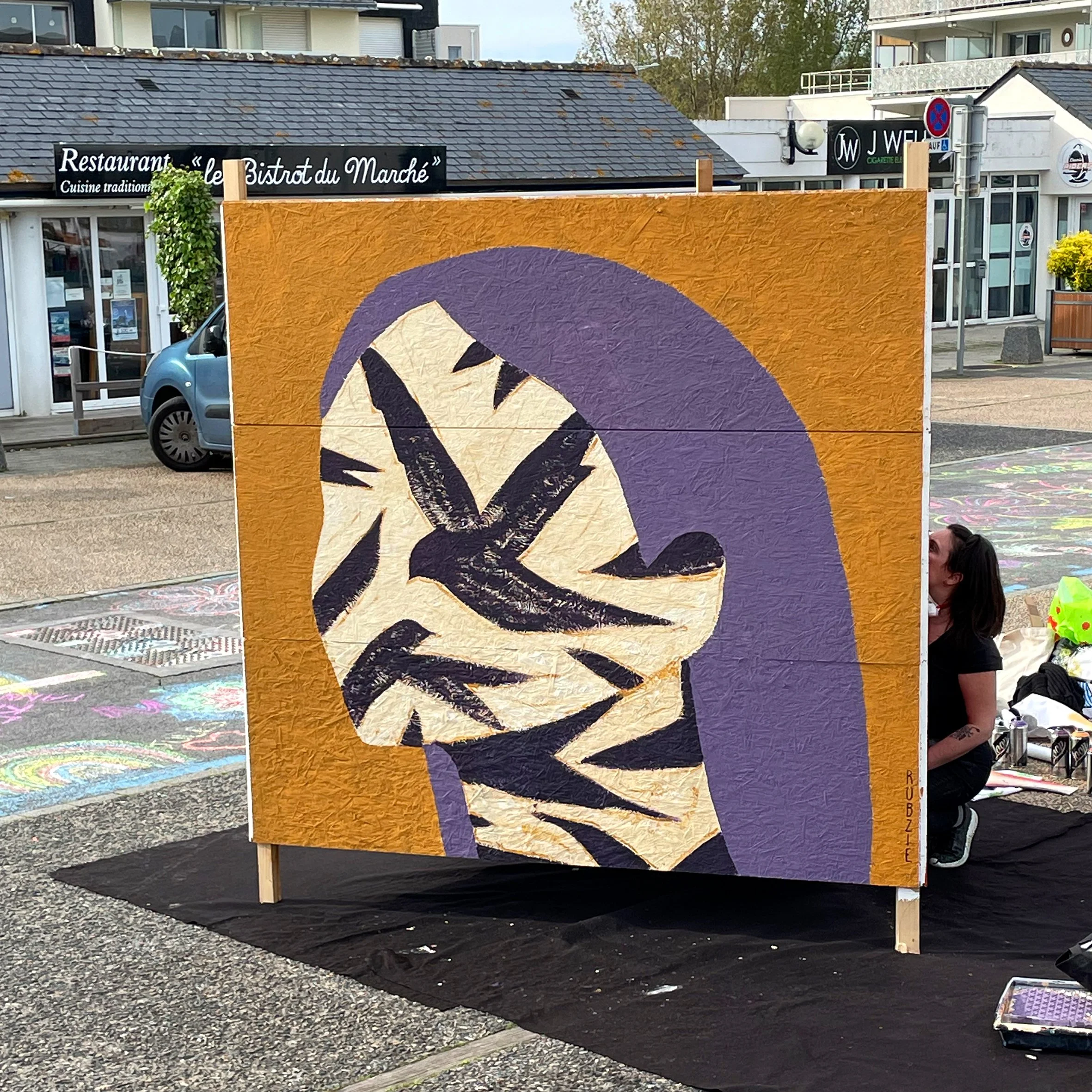 Une fresque artistique représentant un visage stylisé avec des traits abstraits en couleurs violette, blanche et noire, installée dans un espace urbain. Des bâtiments modernes et un véhicule sont visibles en arrière-plan.