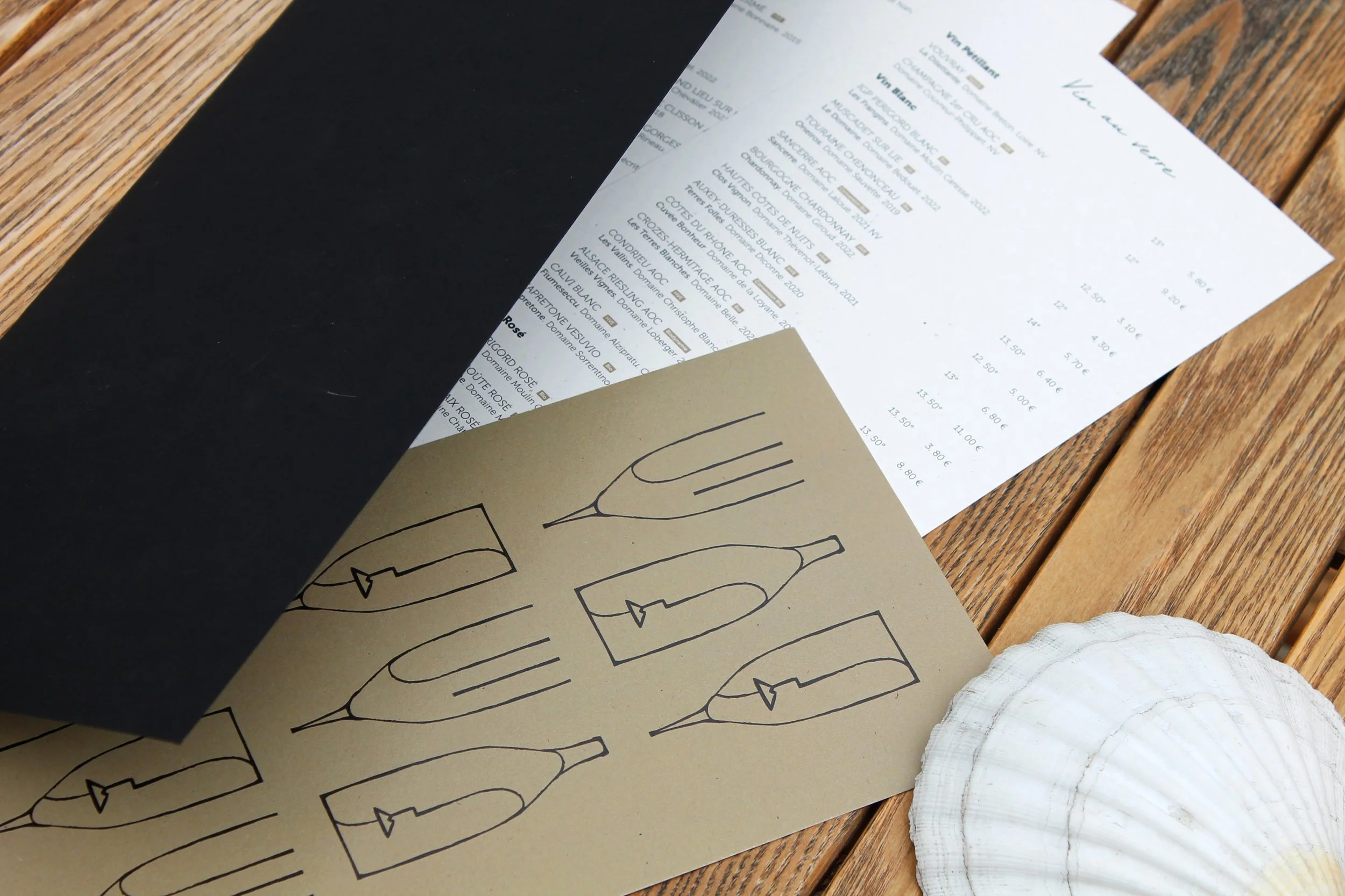 Une fiche de menu de restaurant ou de bar avec une liste de vins et de prix, posée sur une table en bois avec une feuille de papier beige à dessin de bouteilles de vin à côté.