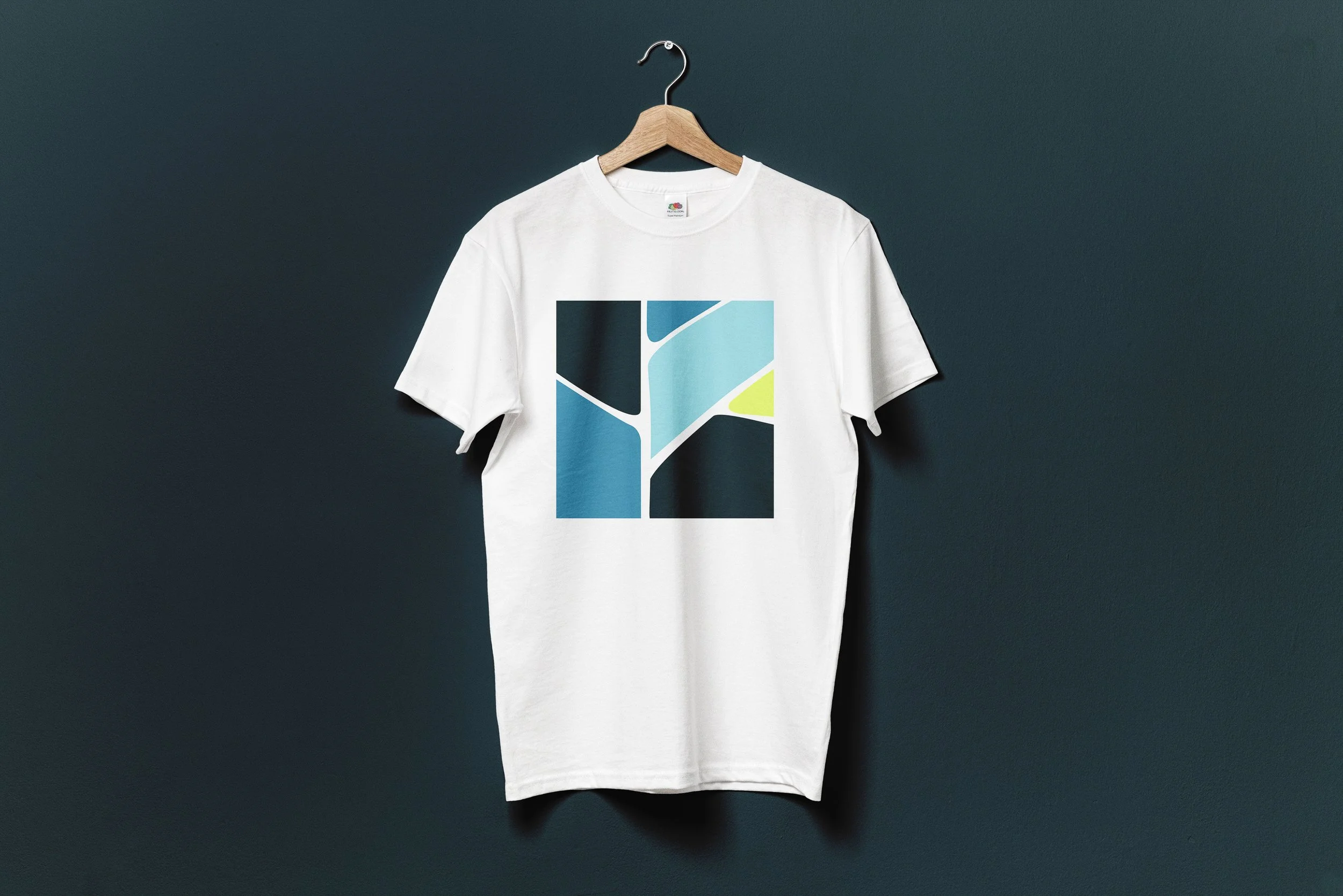 T-shirt blanc avec un design géométrique coloré, suspendu à un cintre en bois accroché à un mur gris foncé.