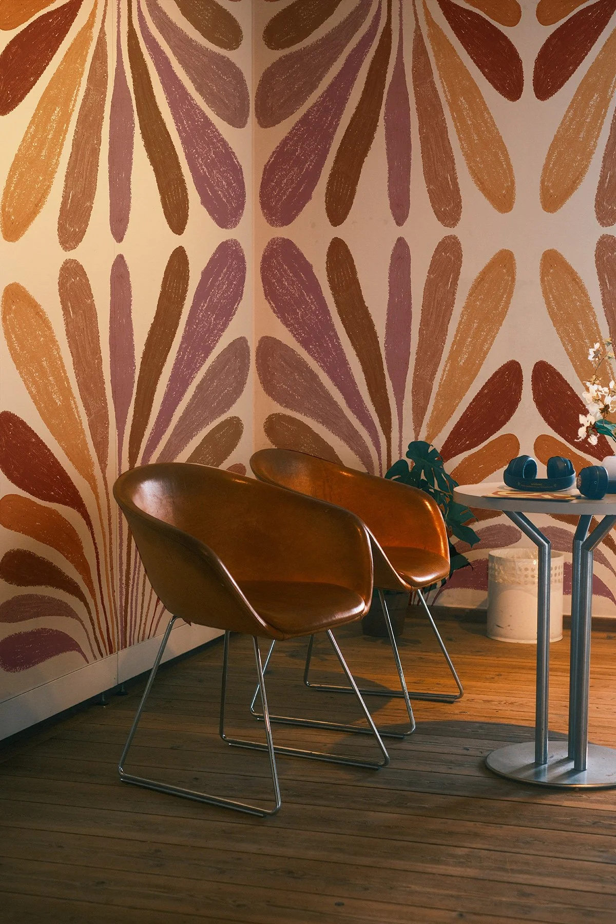 Une pièce avec deux chaises en cuir marron face à un mur avec un motif floral coloré, une petite table blanche avec des écouteurs et une plante verte, un cache-pot blanc et des fleurs blanches.