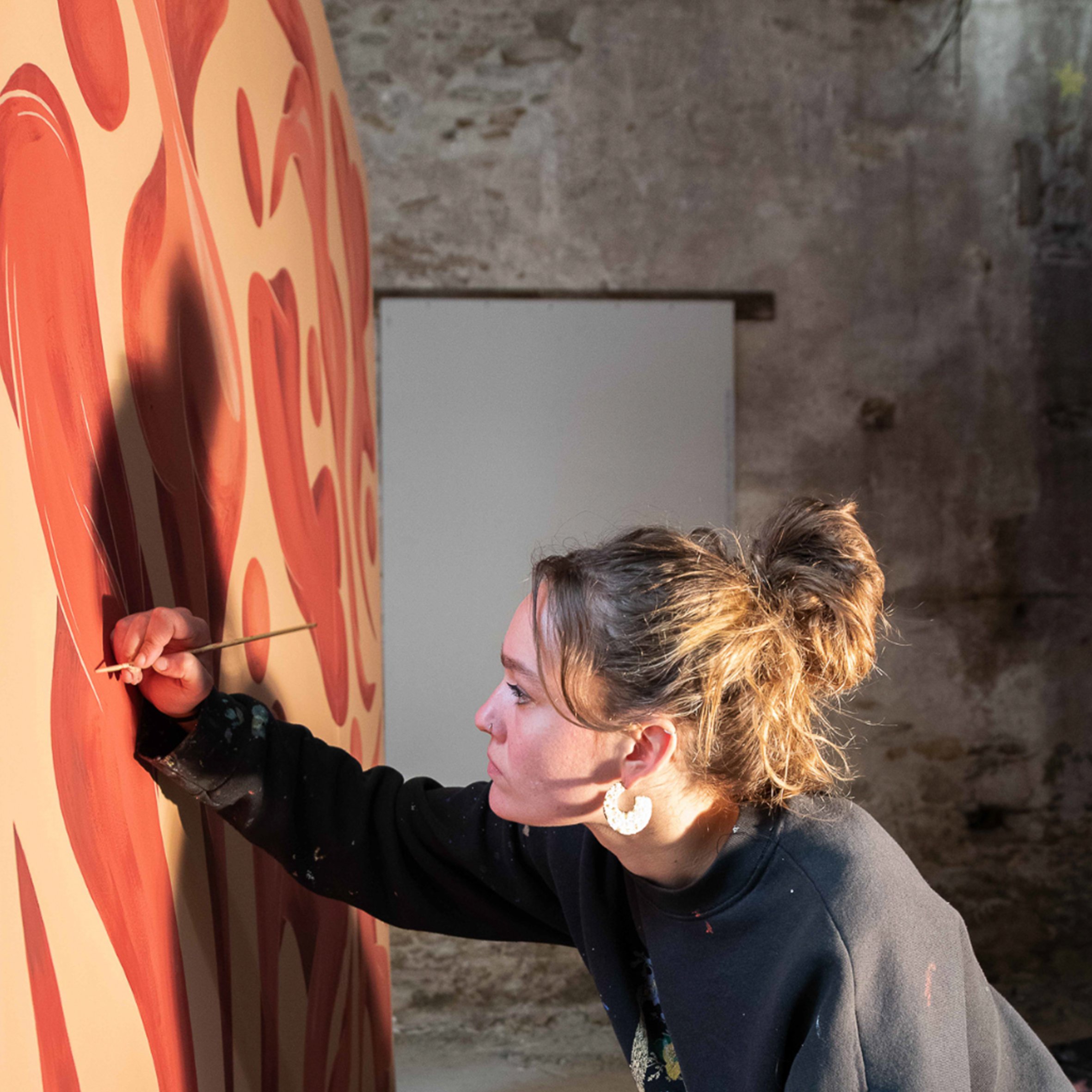 Une femme avec des cheveux bouclés qui se tiens face à une fresque de pizza, tenant un bâton avec une cuillère à la main.