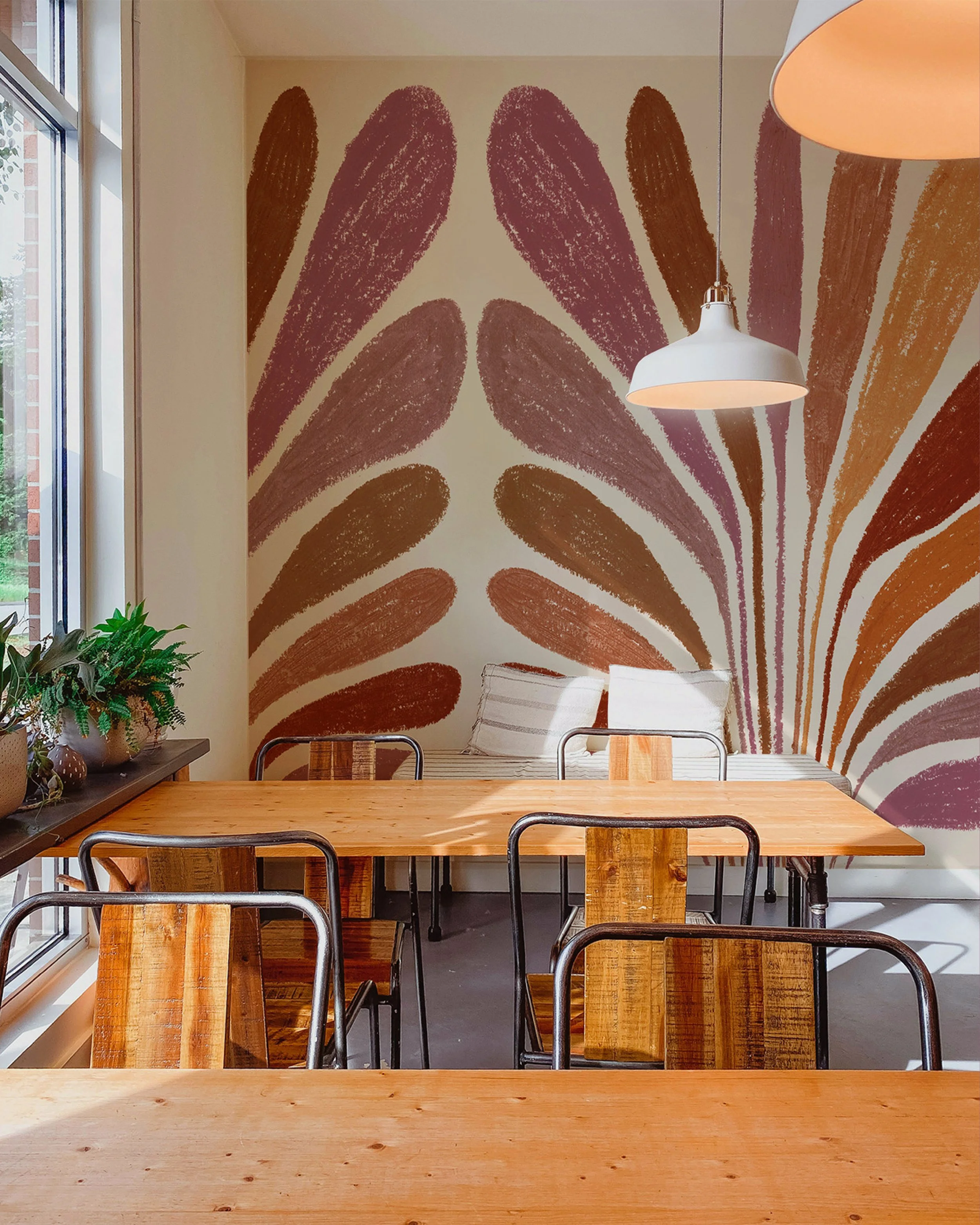 Une salle à manger avec une grande table en bois, des chaises en métal, un mur décoré avec une fresque abstraite aux couleurs marron et beige, et un tapis blanc et beige. Il y a aussi une étagère avec des plantes et des pots à gauche, et une grande fenêtre laissant entrer la lumière naturelle.