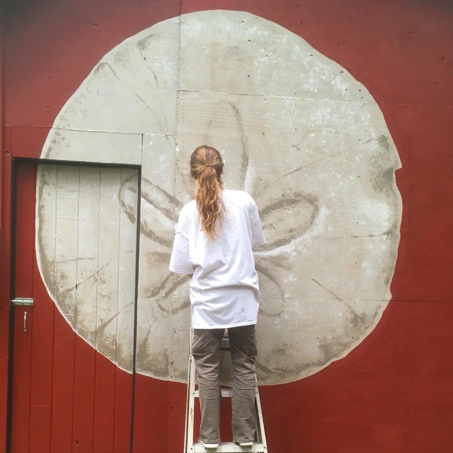 Une personne en t-shirt blanc et pantalon gris se tient sur un escabeau, face à un mur avec une peinture murale représentant un visage en grand format, vu de dos.