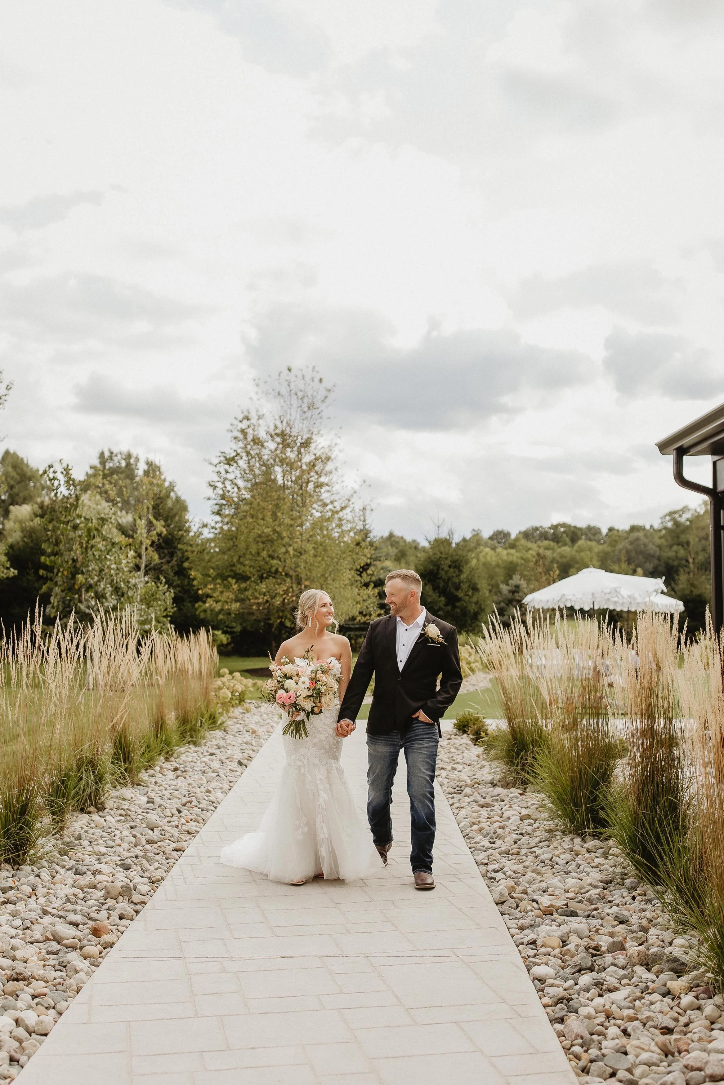 bride and groom holding hands, walking down a pathway with grassy landscaping on either side