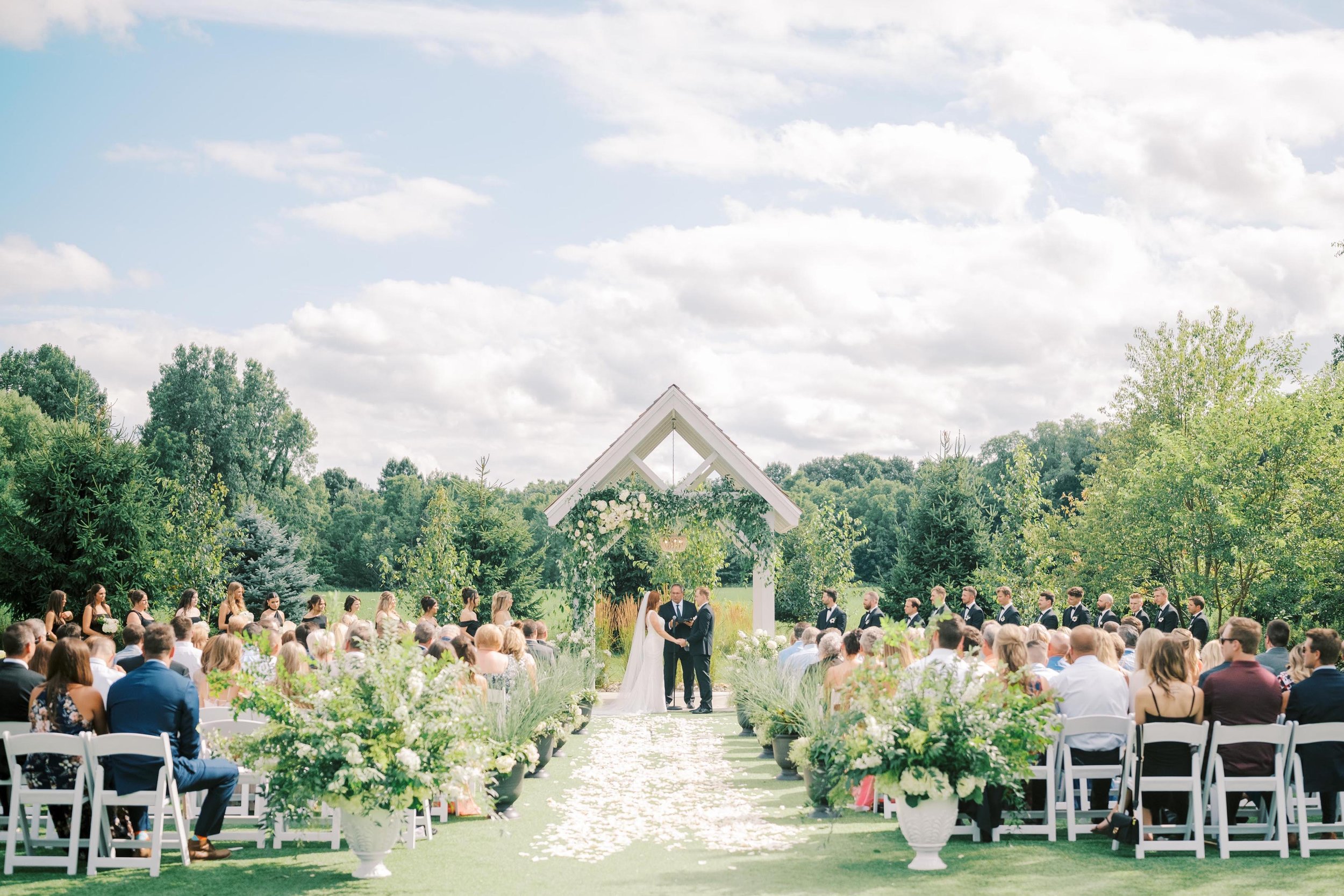 wedding ceremony with rows of guest chairs, the couple standing under the outdoor chapel, and many trees in full summer bloom in the background