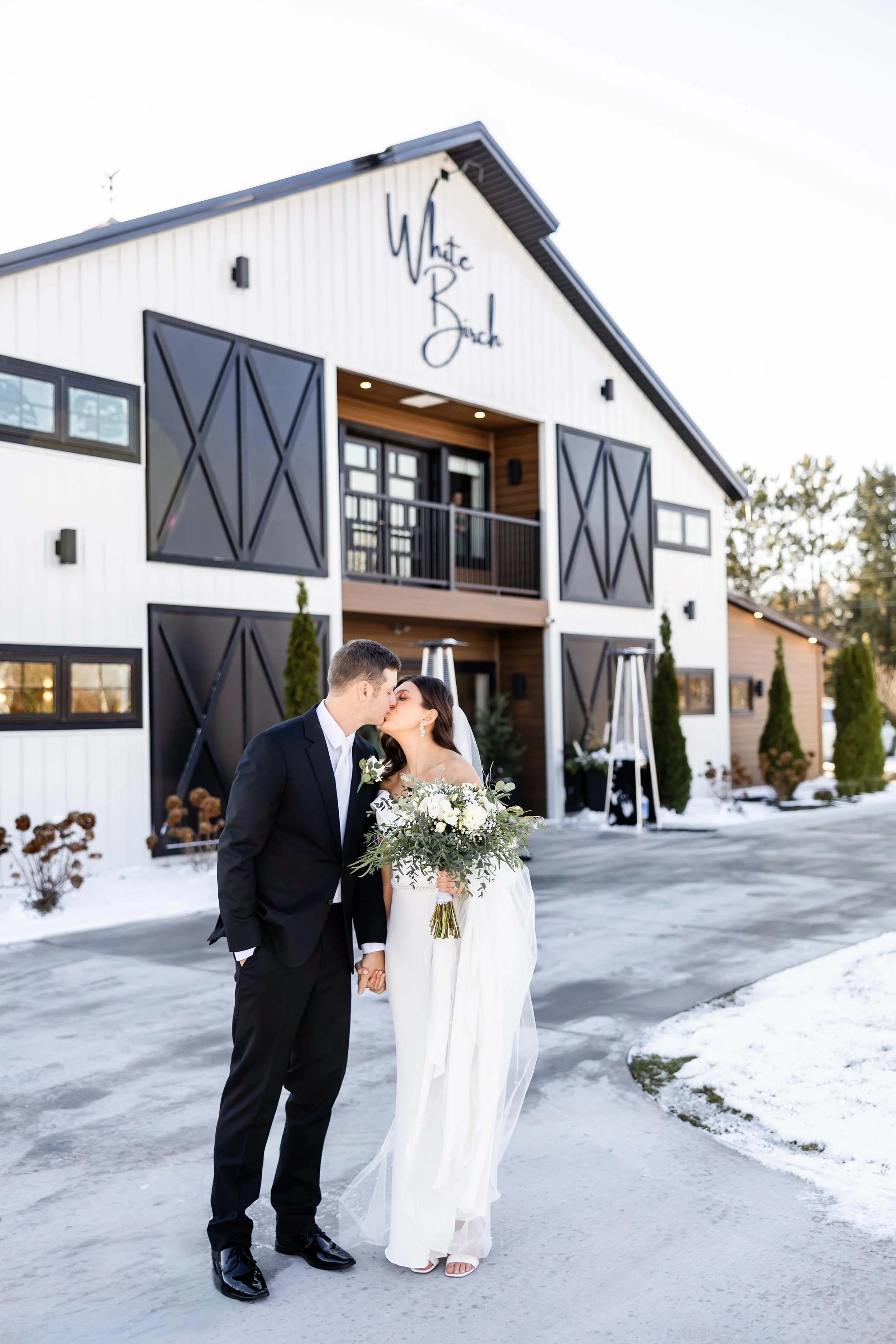couple kissing. on their wedding day in front of White Birch Wedding Barn, with snow covering the landscape