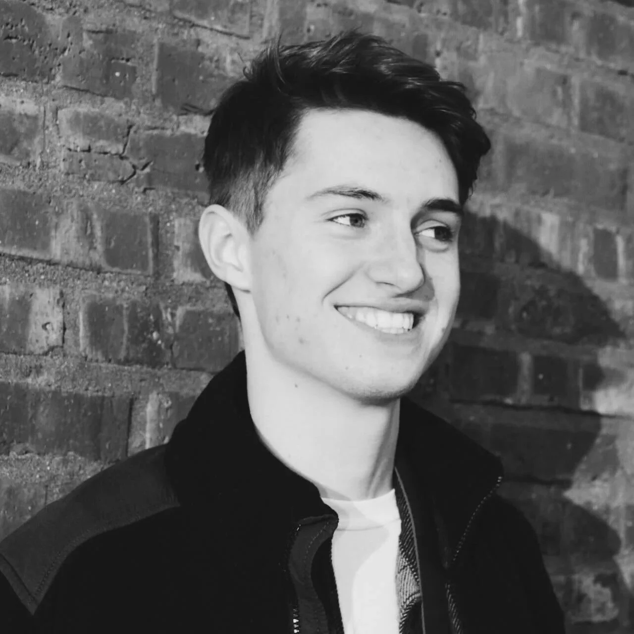 Black and white photo of a young man smiling, standing against a brick wall.