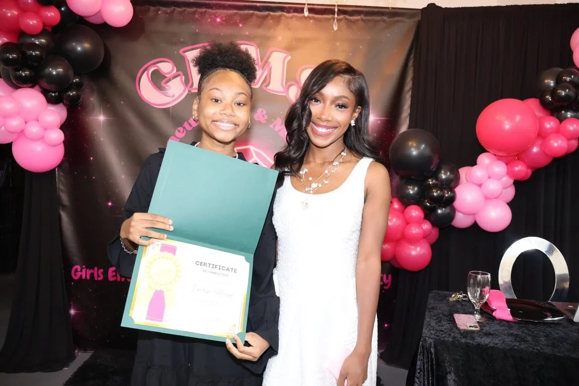 A young girl in black holding a certificate and smiling next to a woman in a white dress, both standing in front of a backdrop with pink and black balloons at a girls event.