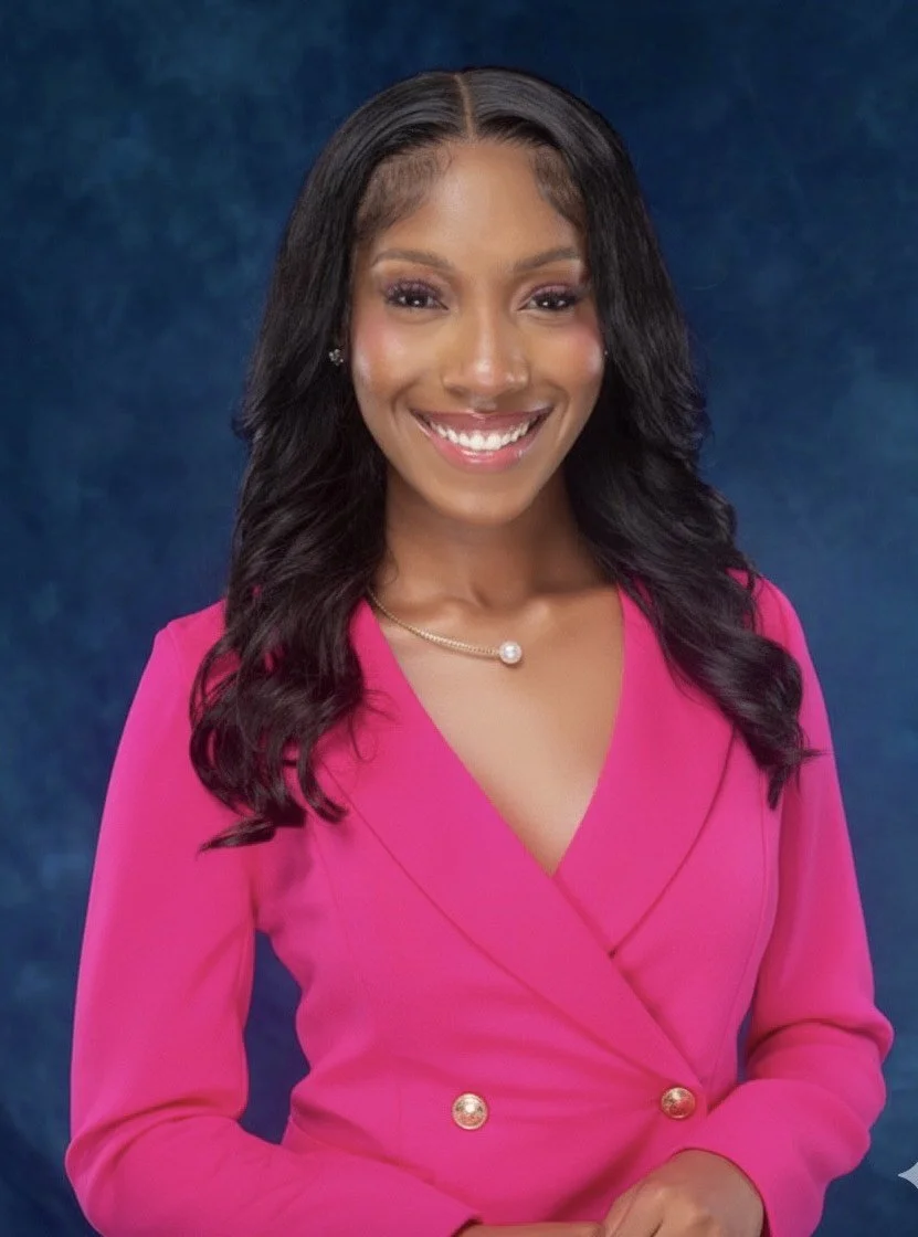 A smiling woman with long, wavy black hair, wearing a bright pink blazer with gold buttons and a pearl necklace, against a dark blue background.
