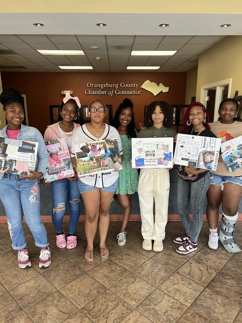 Seven young women standing indoors in front of a wall that reads 'Orangeburg County Chamber of Commerce,' holding posters and smiling at the camera.