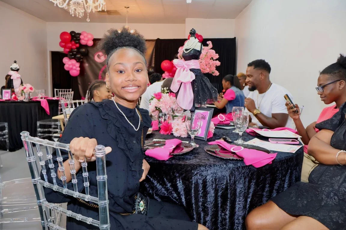 A young girl sitting at a decorated table with pink and black accents, celebrating a special occasion, likely a birthday or party, with other guests and festive decorations in the background.