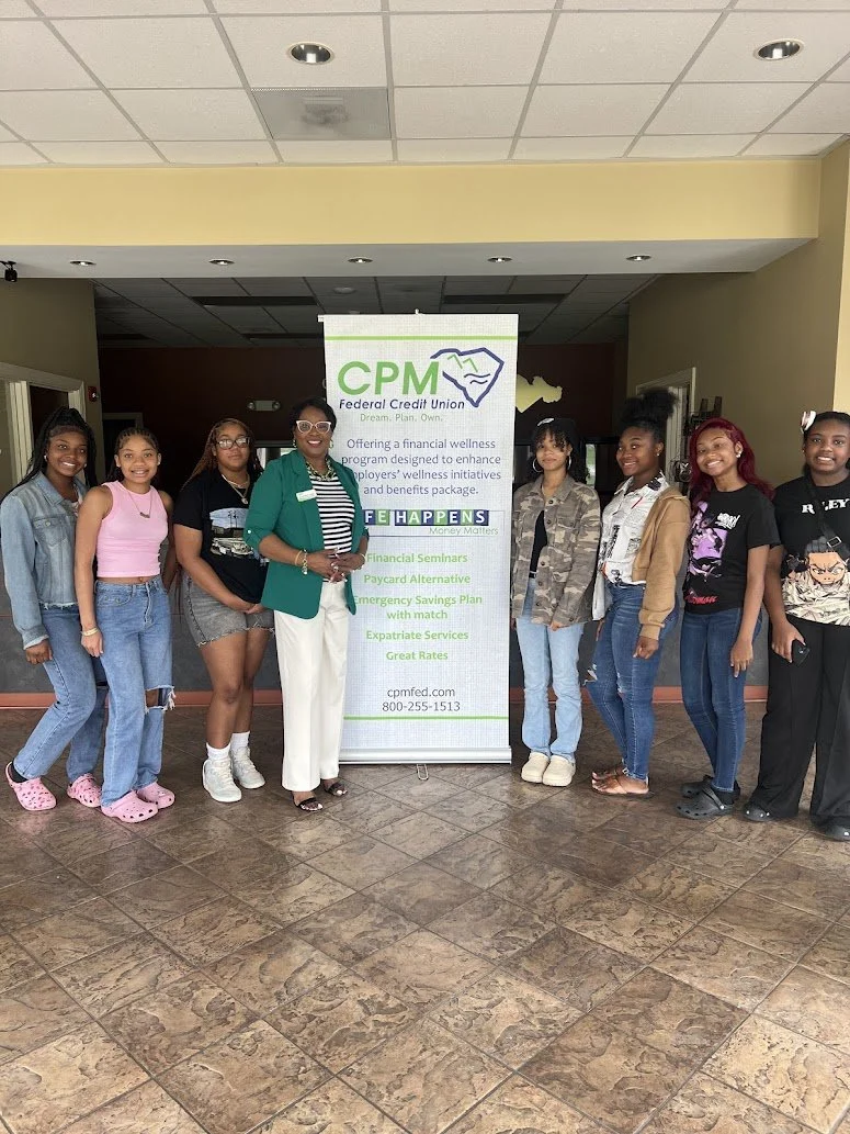 Group of eight women standing next to a CPM Federal Credit Union promotional banner in a lobby. They are smiling and posing for the photo.